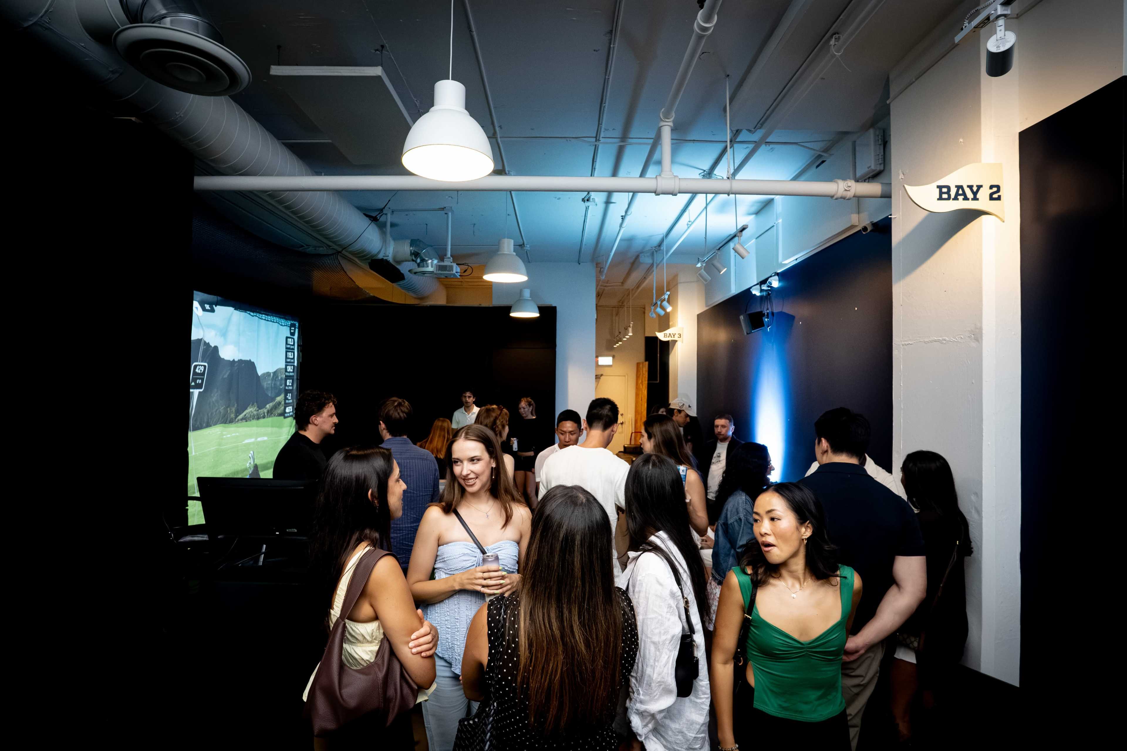 A group of people mingles in a spacious indoor setting, with a screen displaying an image in the background.