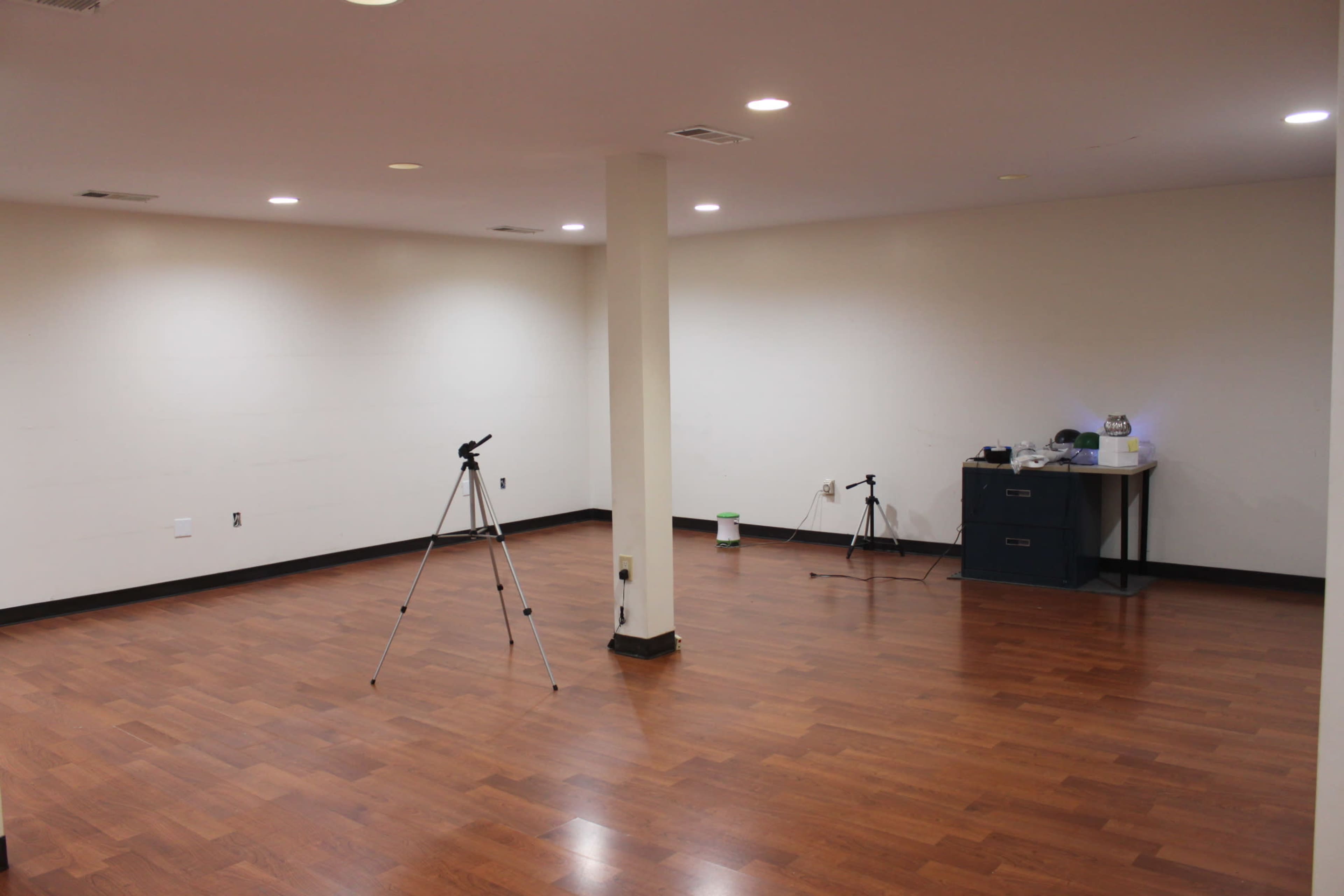 The image shows an empty room with hardwood floors, a few tripods set up, and a table in the corner holding equipment.