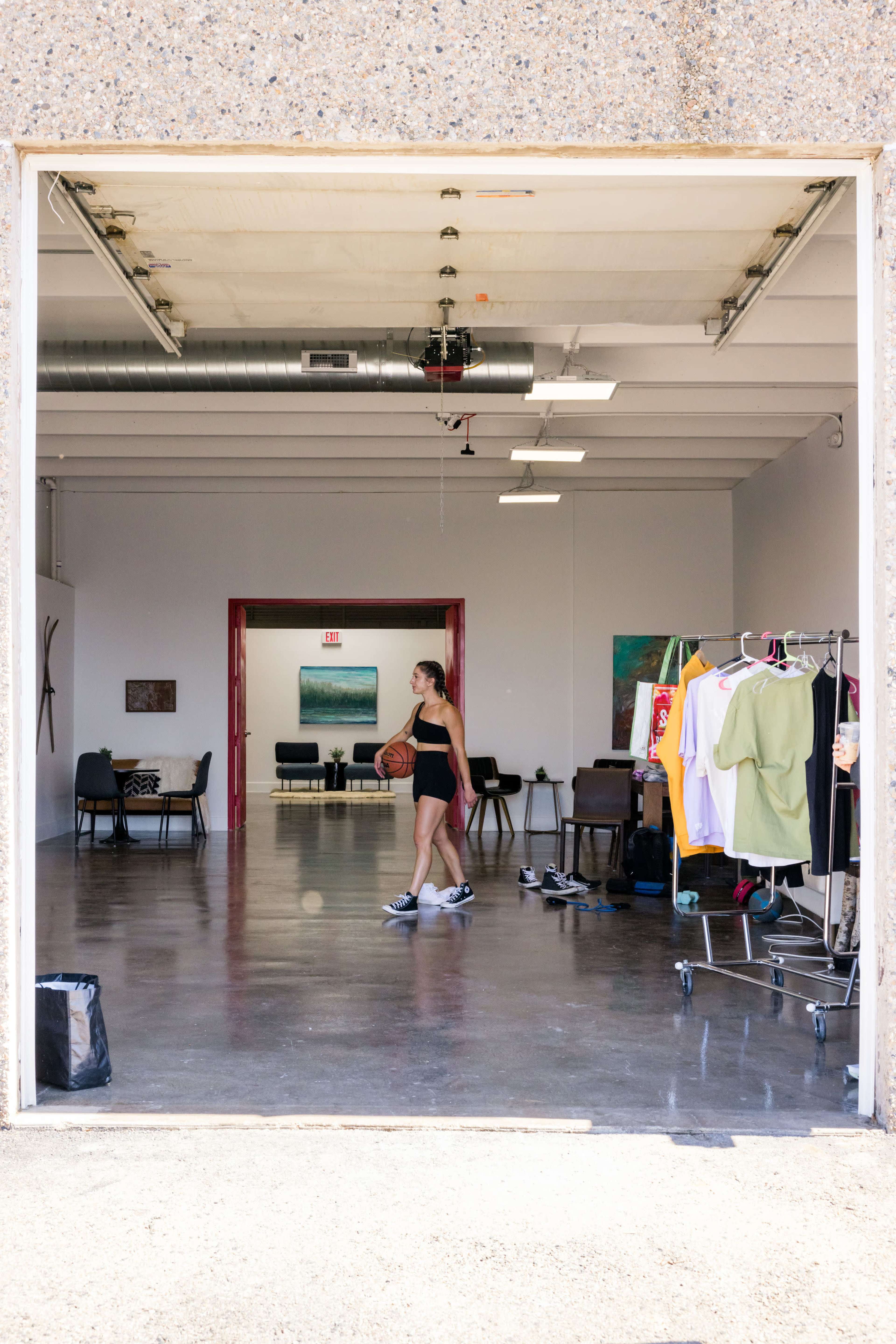A woman in athletic wear holds a basketball while walking through an open doorway of a spacious room filled with chairs and clothing racks.