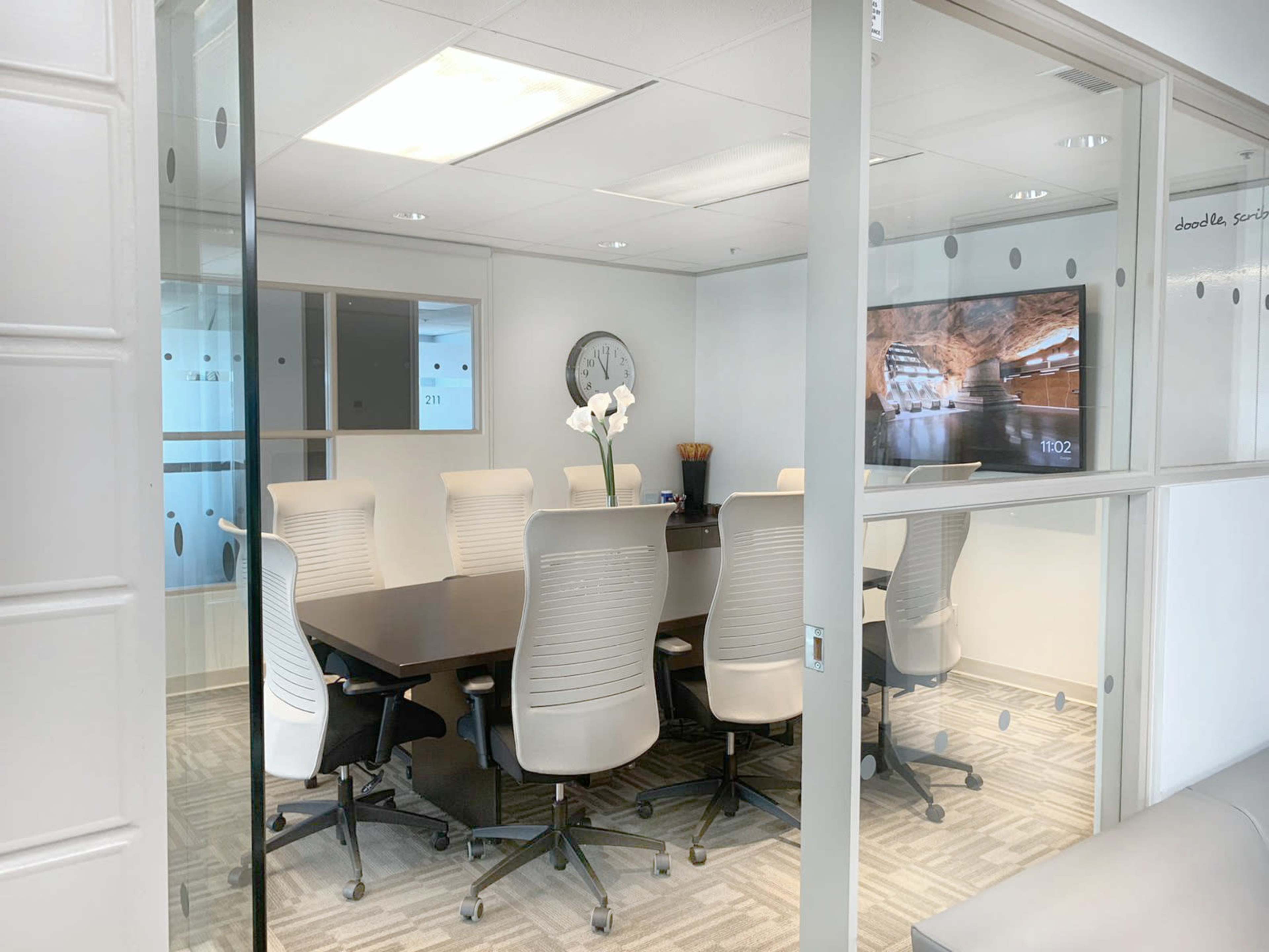A glass-walled conference room features a large table surrounded by ergonomic chairs and a television displaying a scenic image.