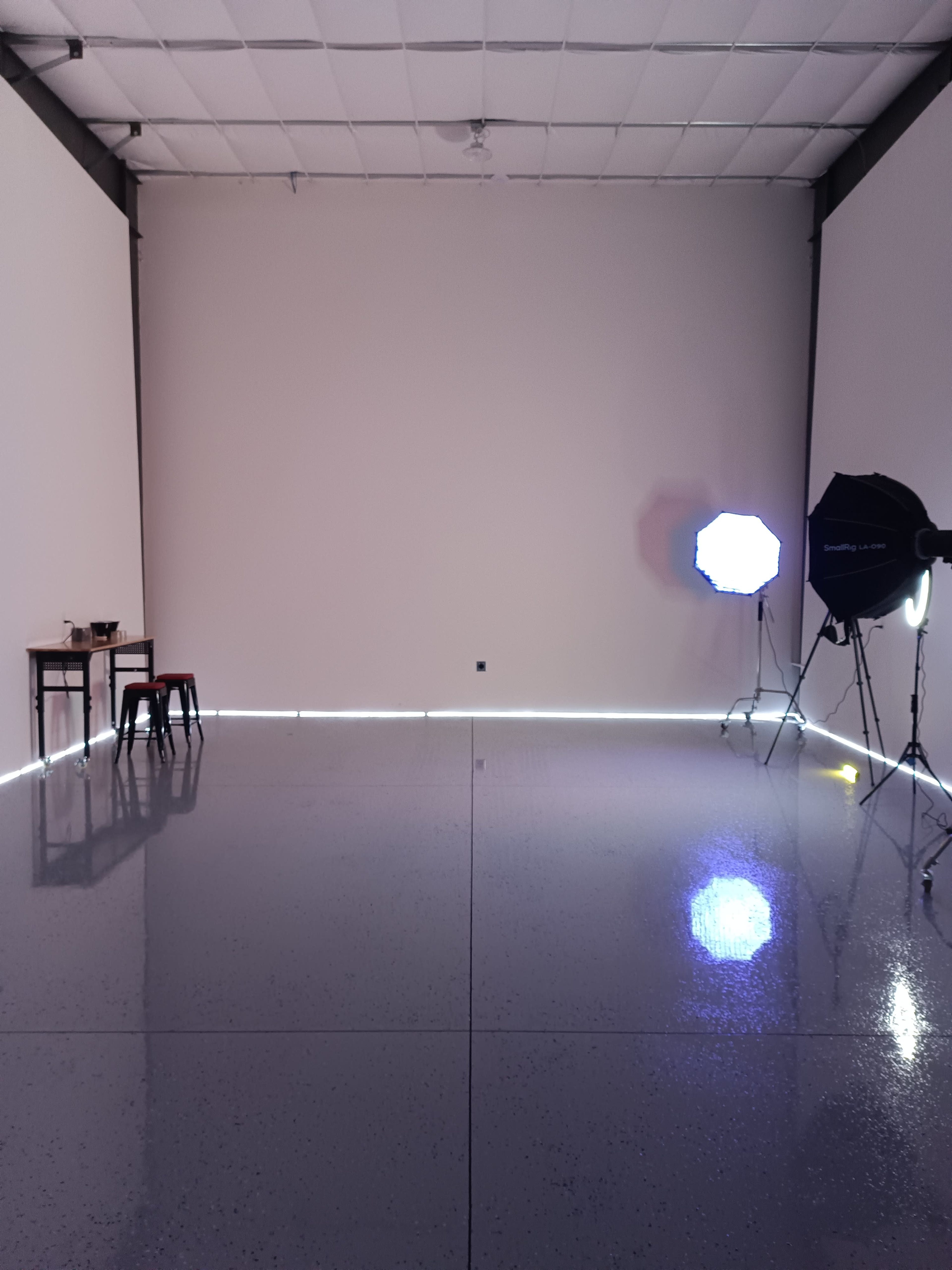 The image shows a blank, well-lit studio space featuring a table and two stools on one side and lighting equipment on the other.