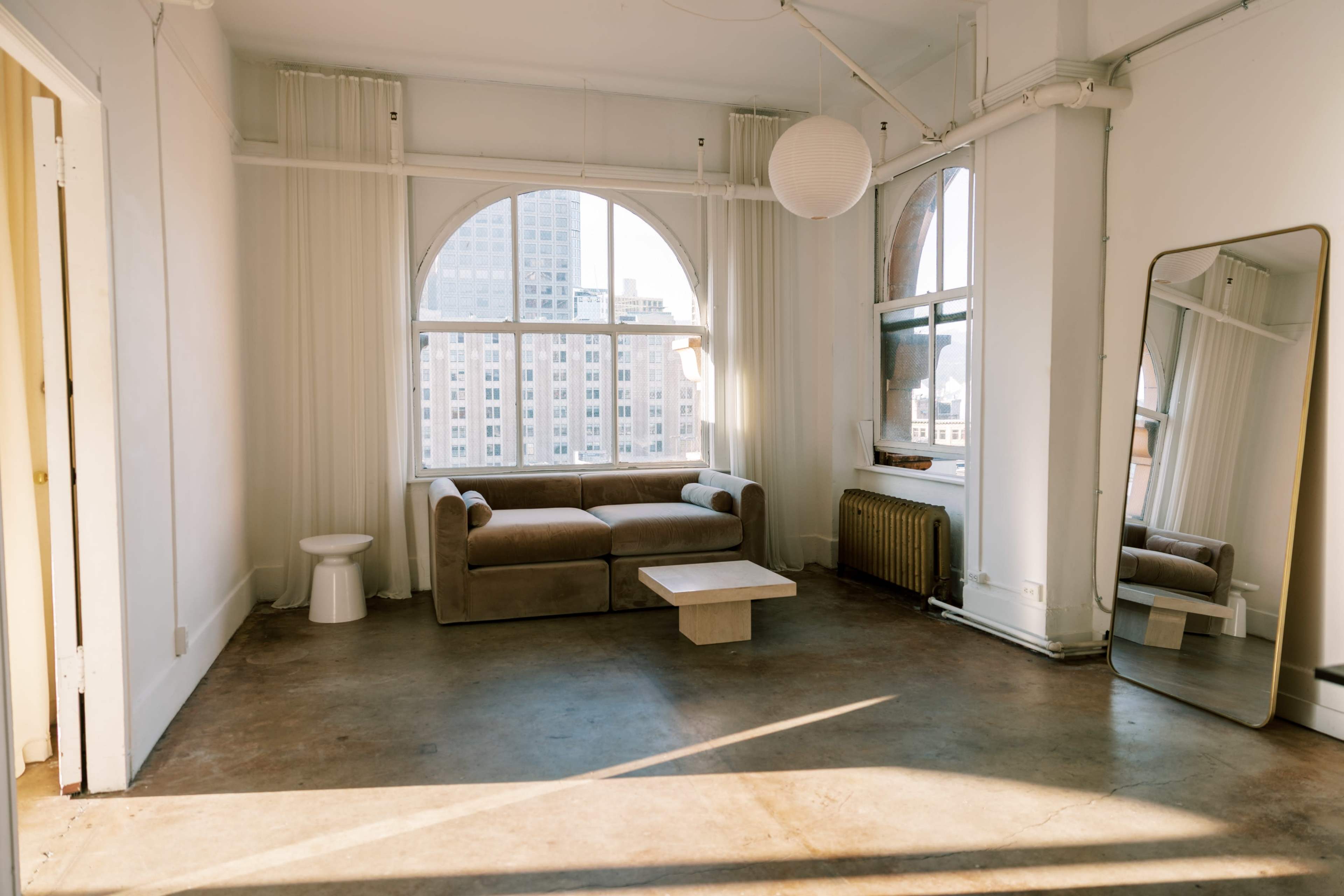 The image shows a spacious, well-lit living area featuring a beige sofa, a small white table, a large mirror, and a window with city views.