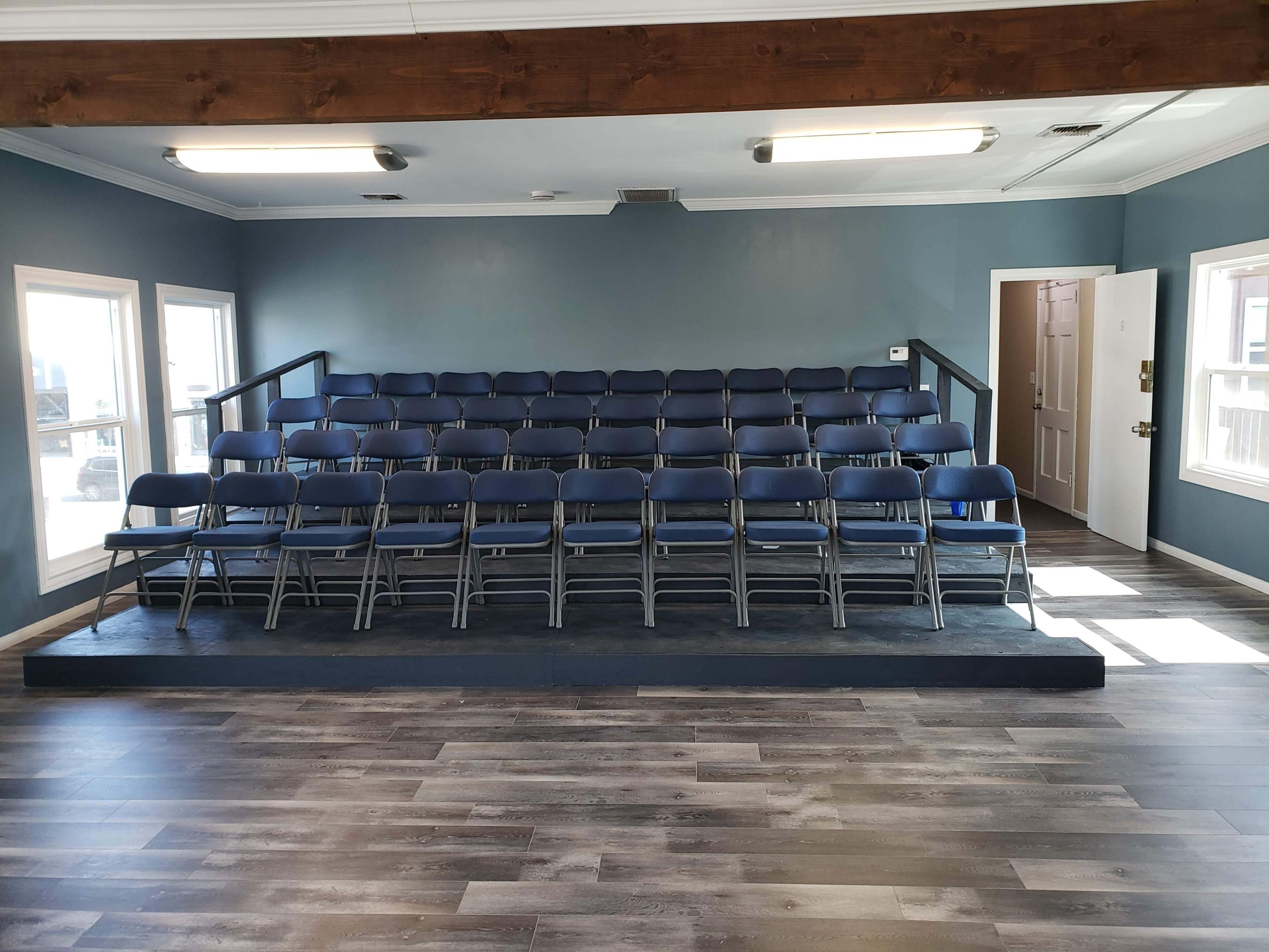 A small auditorium with stacked blue folding chairs arranged in a tiered seating layout and a wooden beam along the ceiling.
