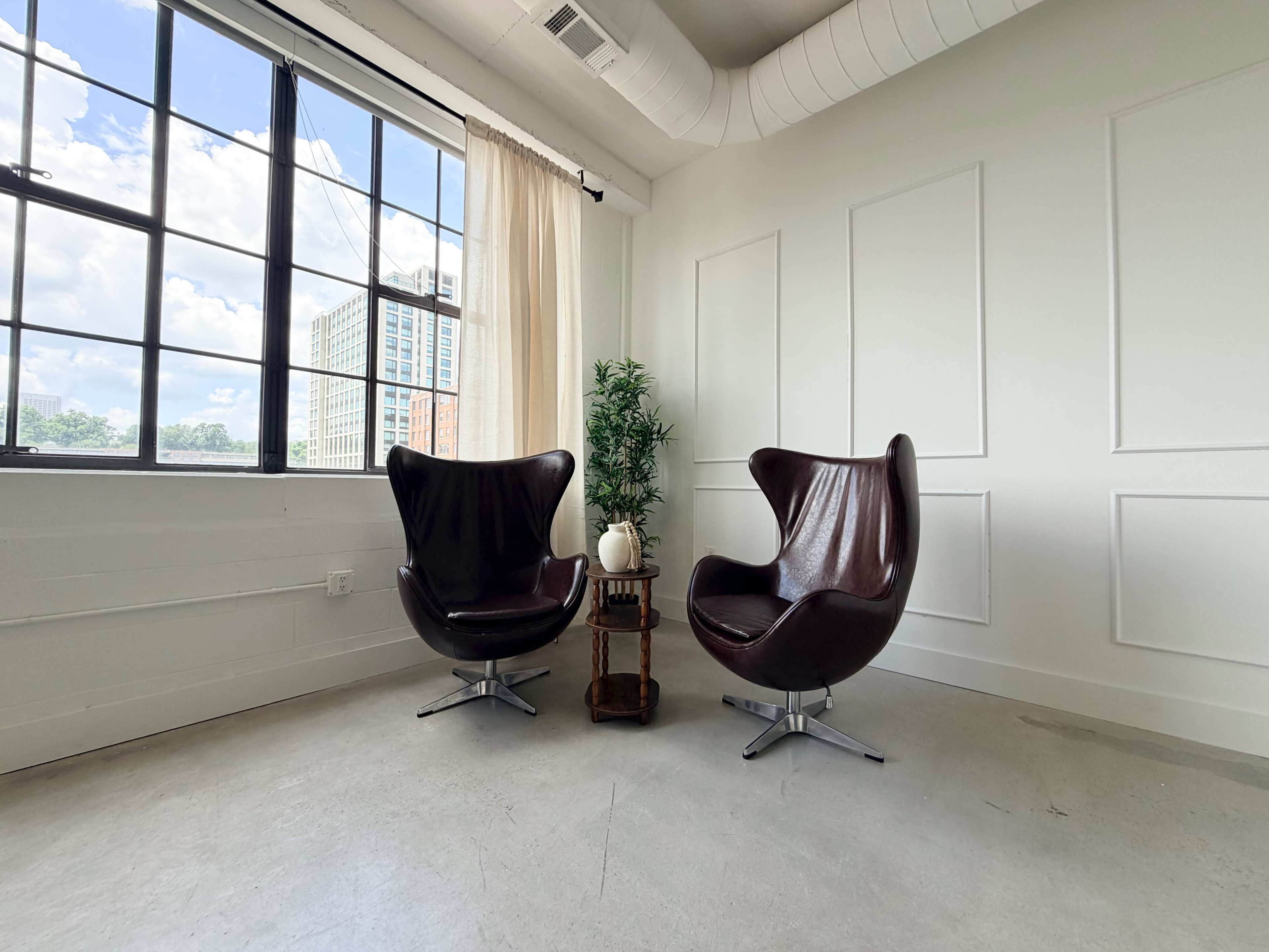 The image shows two brown modern chairs facing each other near a window, with a small table and a potted plant between them.