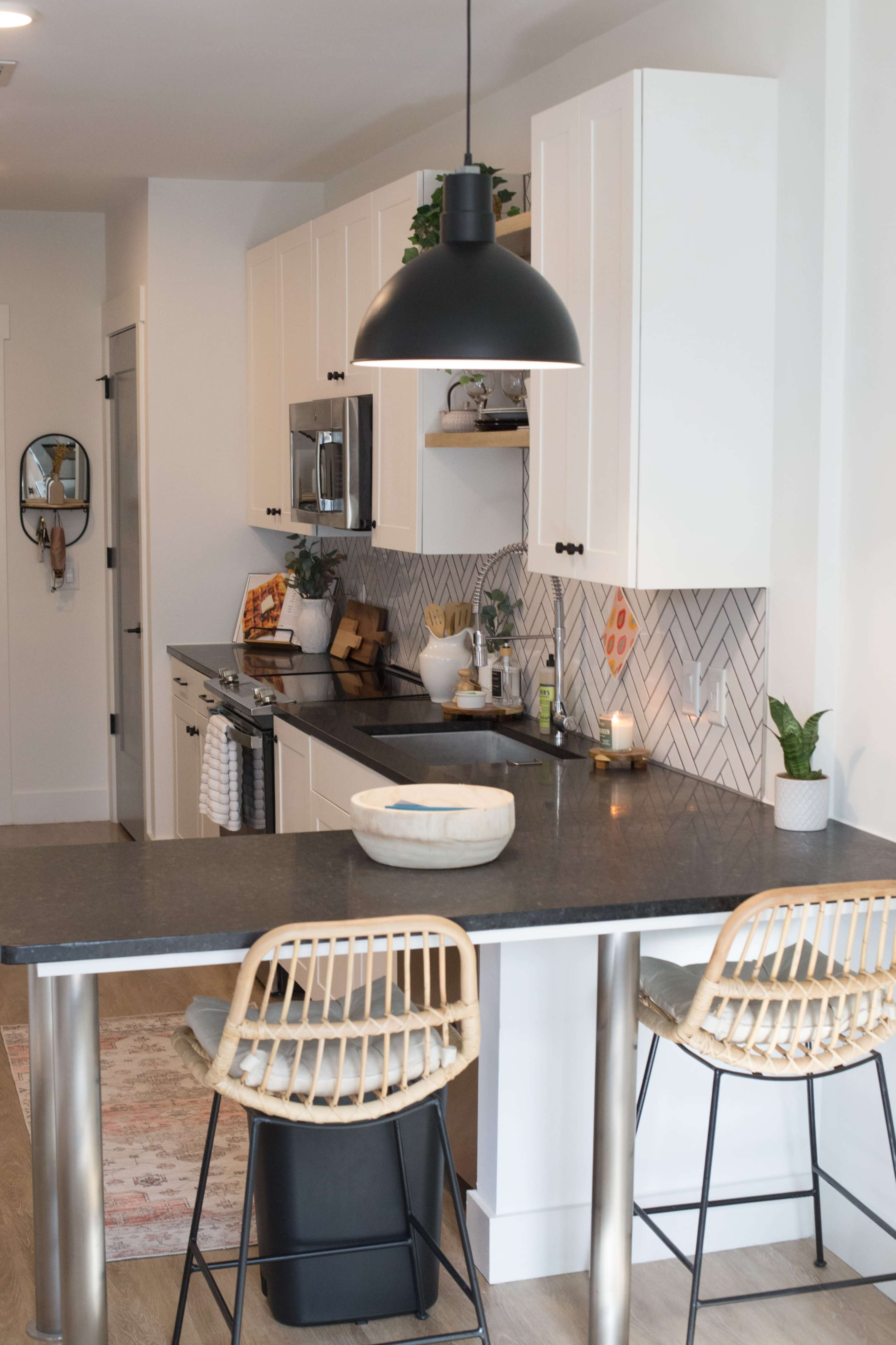A modern kitchen features white cabinetry, a black countertop, and a herringbone tile backsplash, with two woven bar stools at a small breakfast bar.