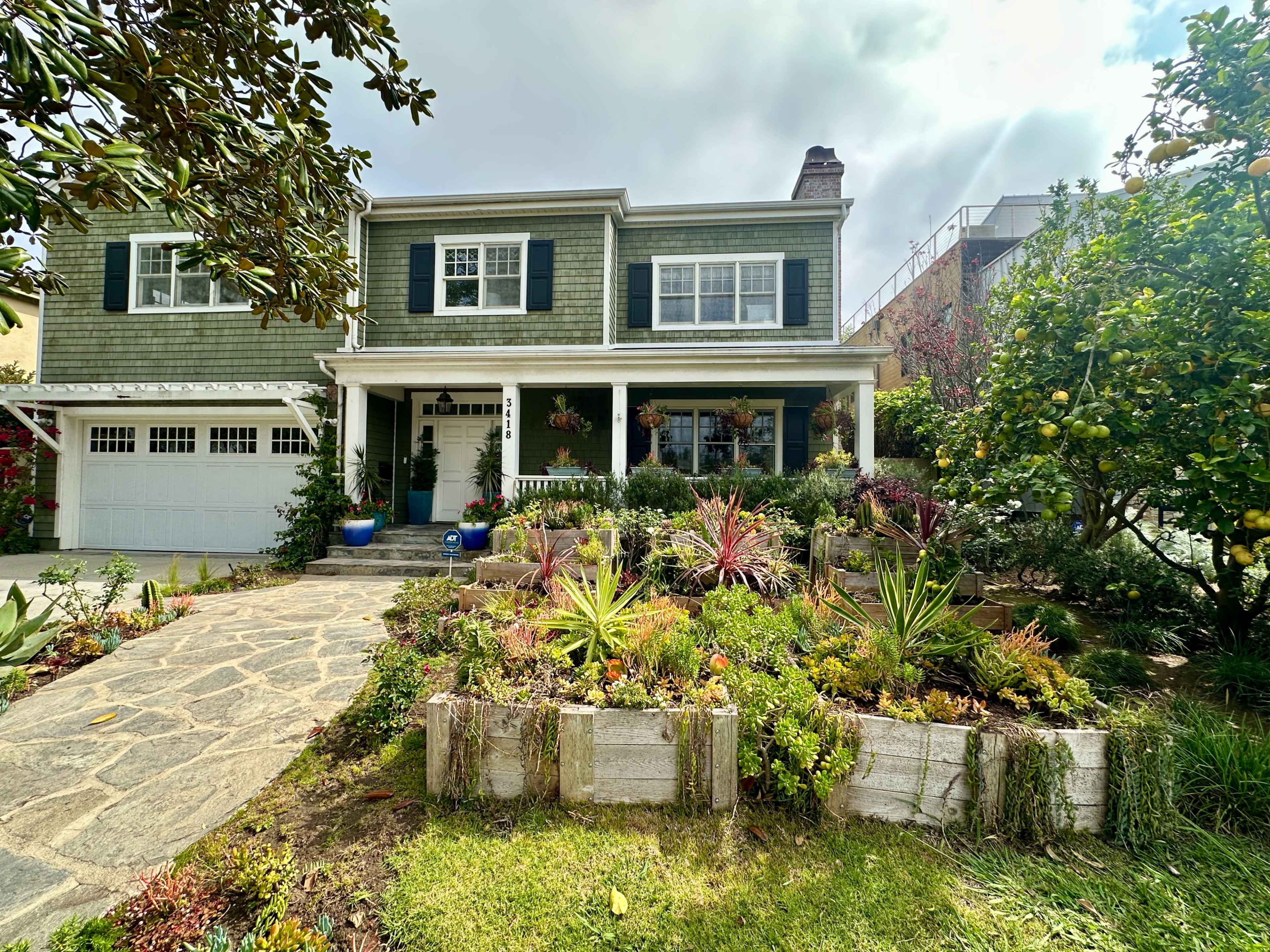 A two-story green house with white trim features a landscaped front yard filled with various plants and stone pathways.