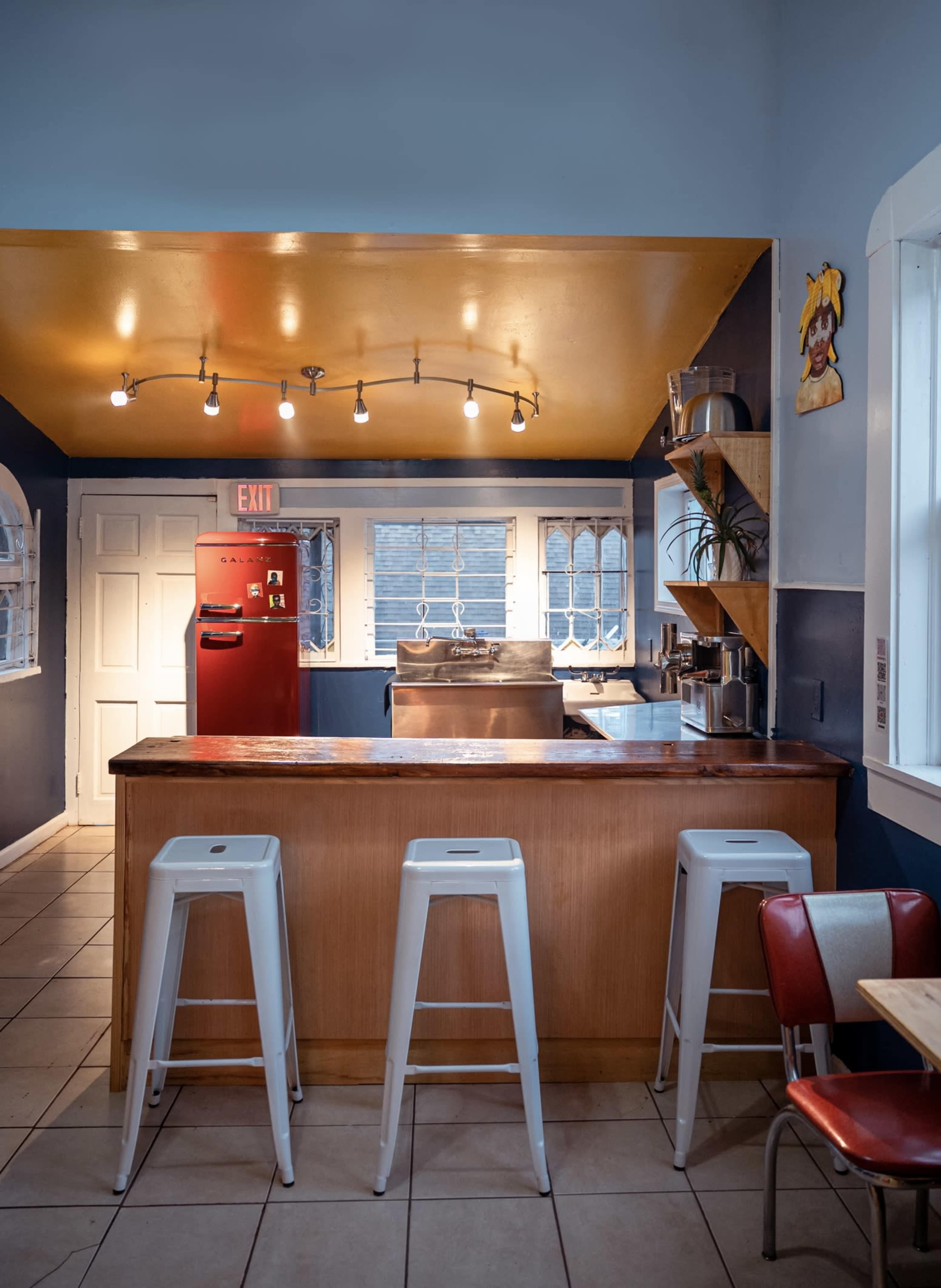 A modern kitchen with a wooden bar, a red refrigerator, metal stools, and bright yellow and blue walls.