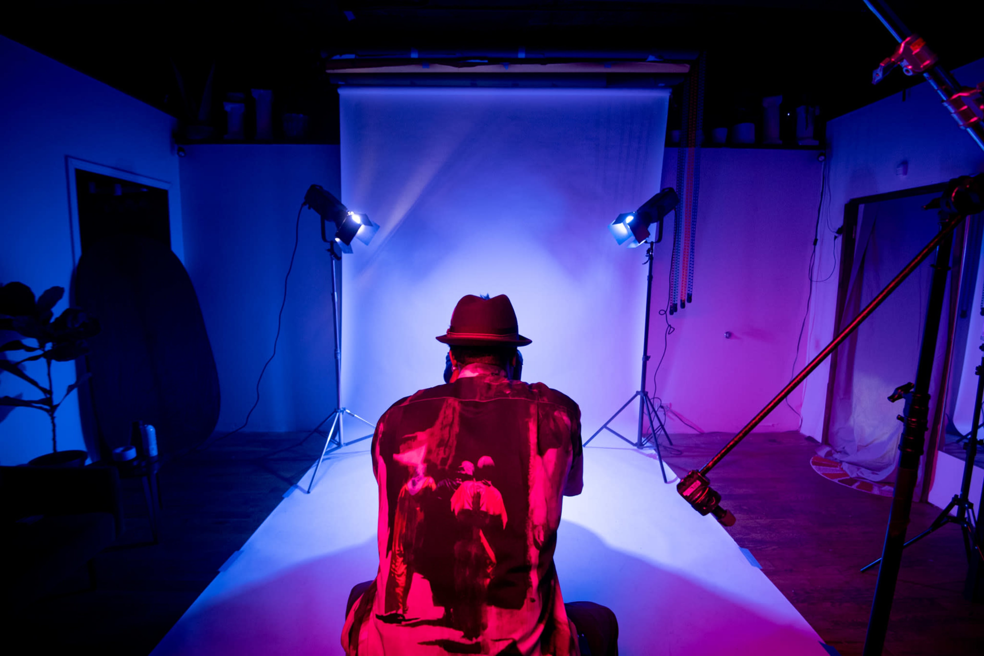 A person wearing a hat sits on the floor in a photography studio, facing a backdrop illuminated by colorful lights.