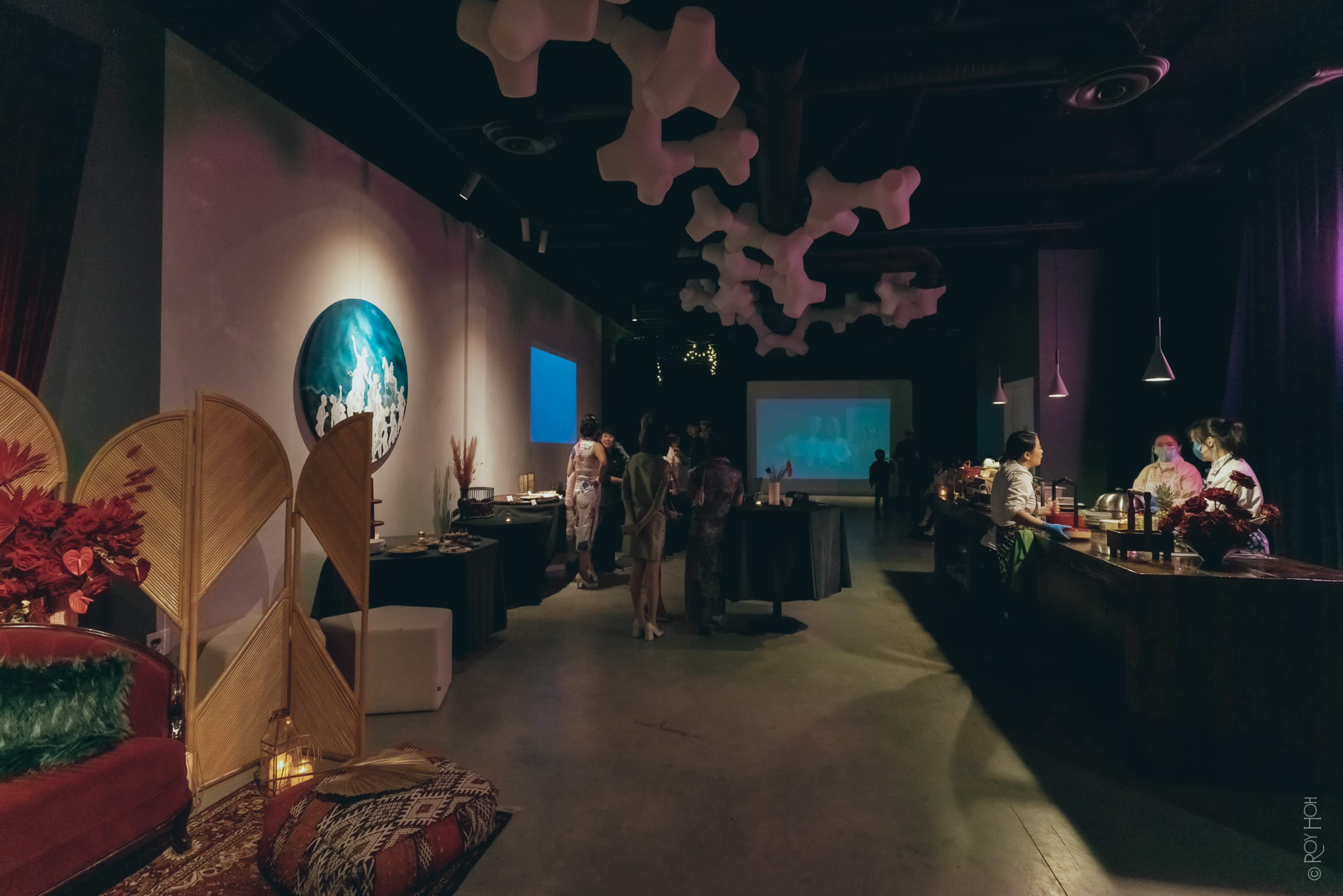 A spacious event room features a bar, dining tables, and decorative elements, with guests mingling and artworks displayed on the walls.