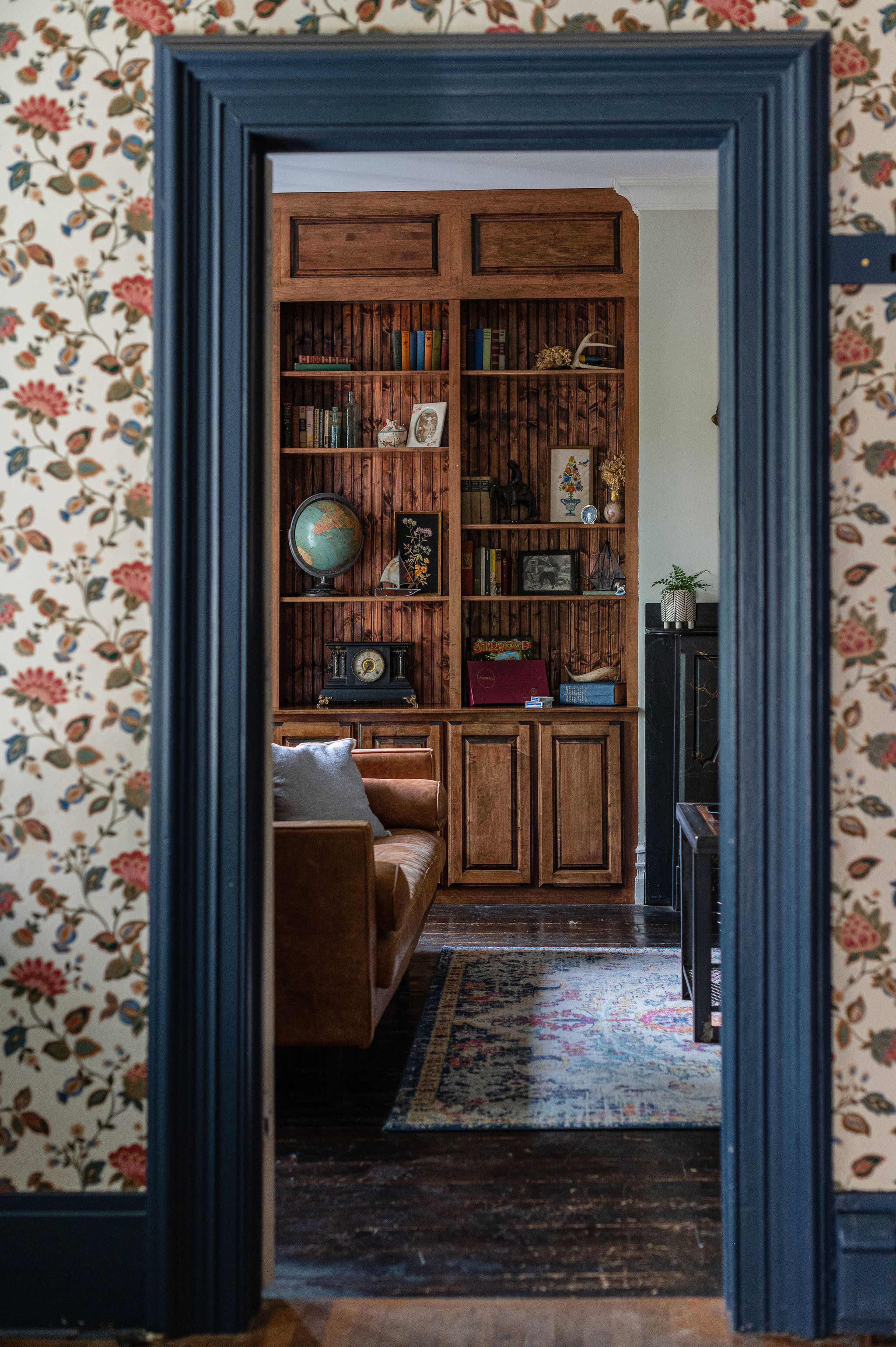 A wooden bookshelf filled with books and decorative items is visible through a doorway leading to a cozy room with floral wallpaper and a leather couch.