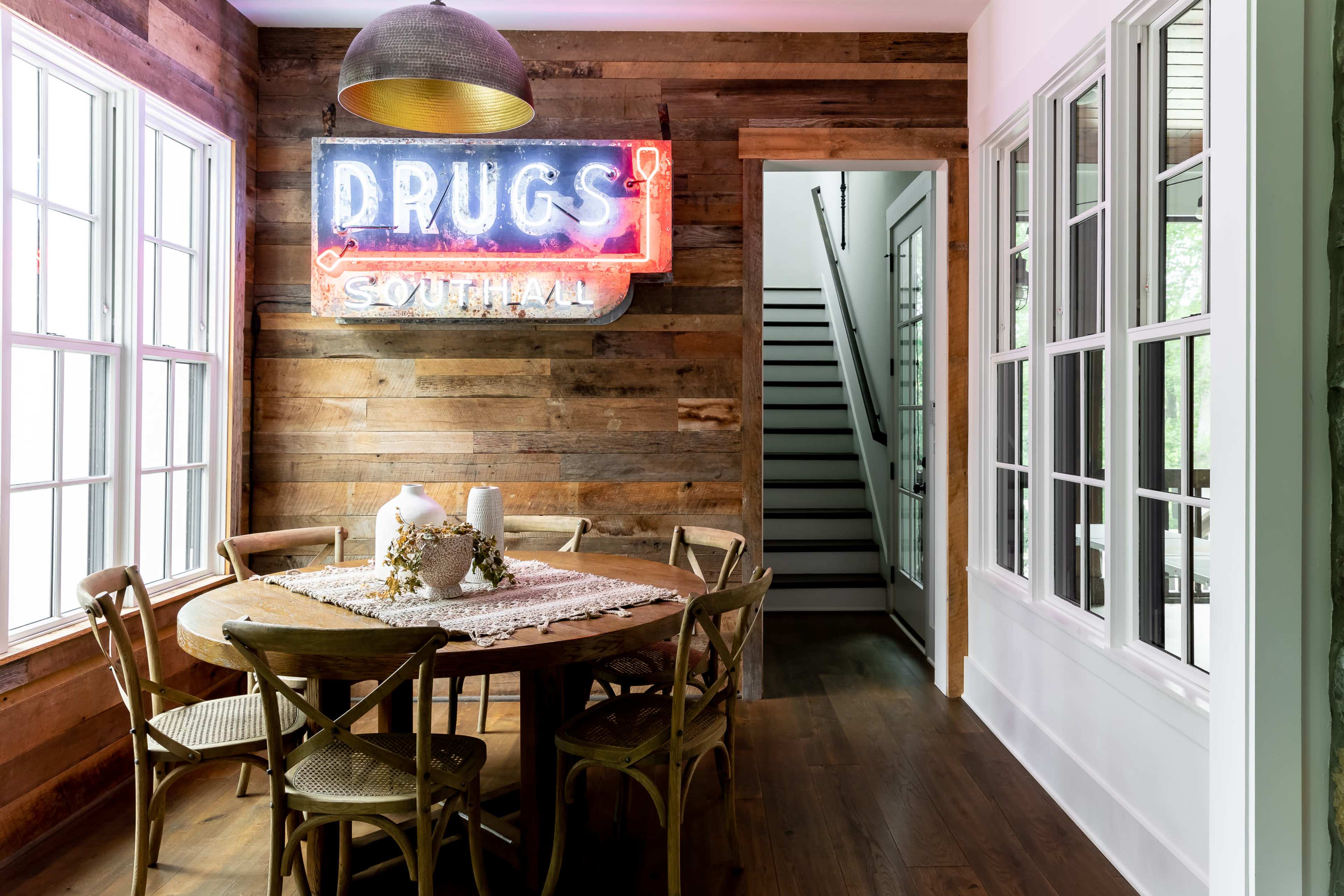 A dining area features a wooden table surrounded by chairs, with a neon sign reading "DRUGS SOUTH" on a rustic wooden wall and a staircase visible in the background.
