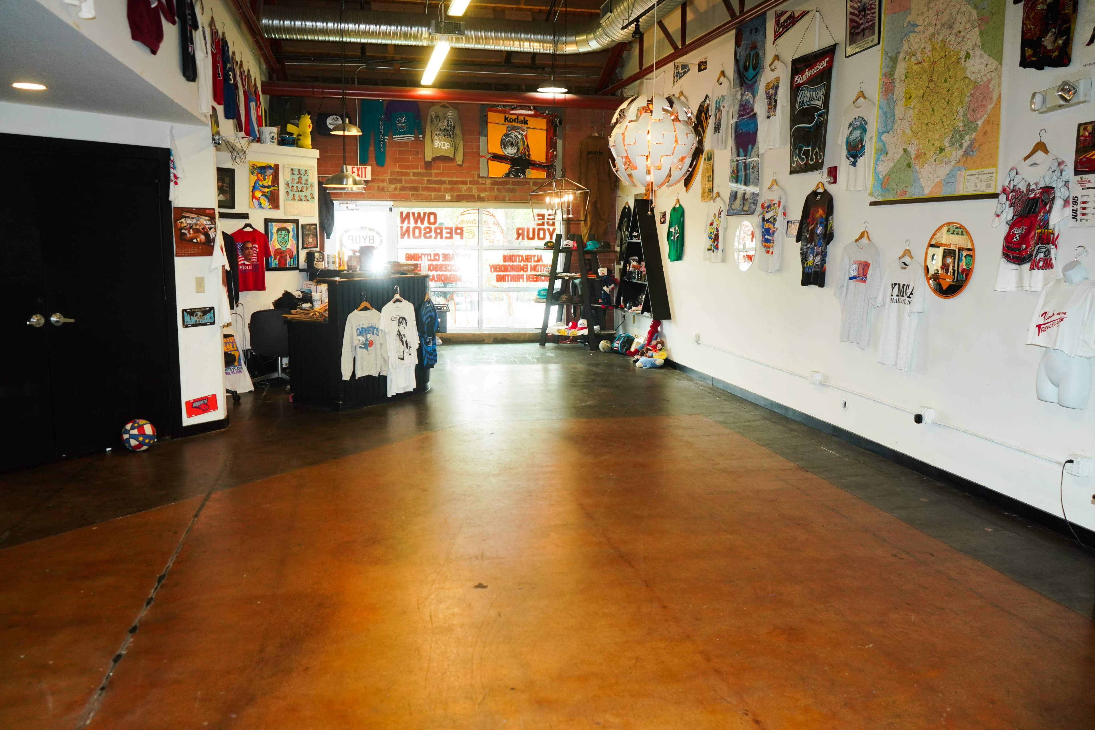 An empty clothing store interior with a polished concrete floor, walls adorned with various shirts and posters, and a large window displaying promotional text.