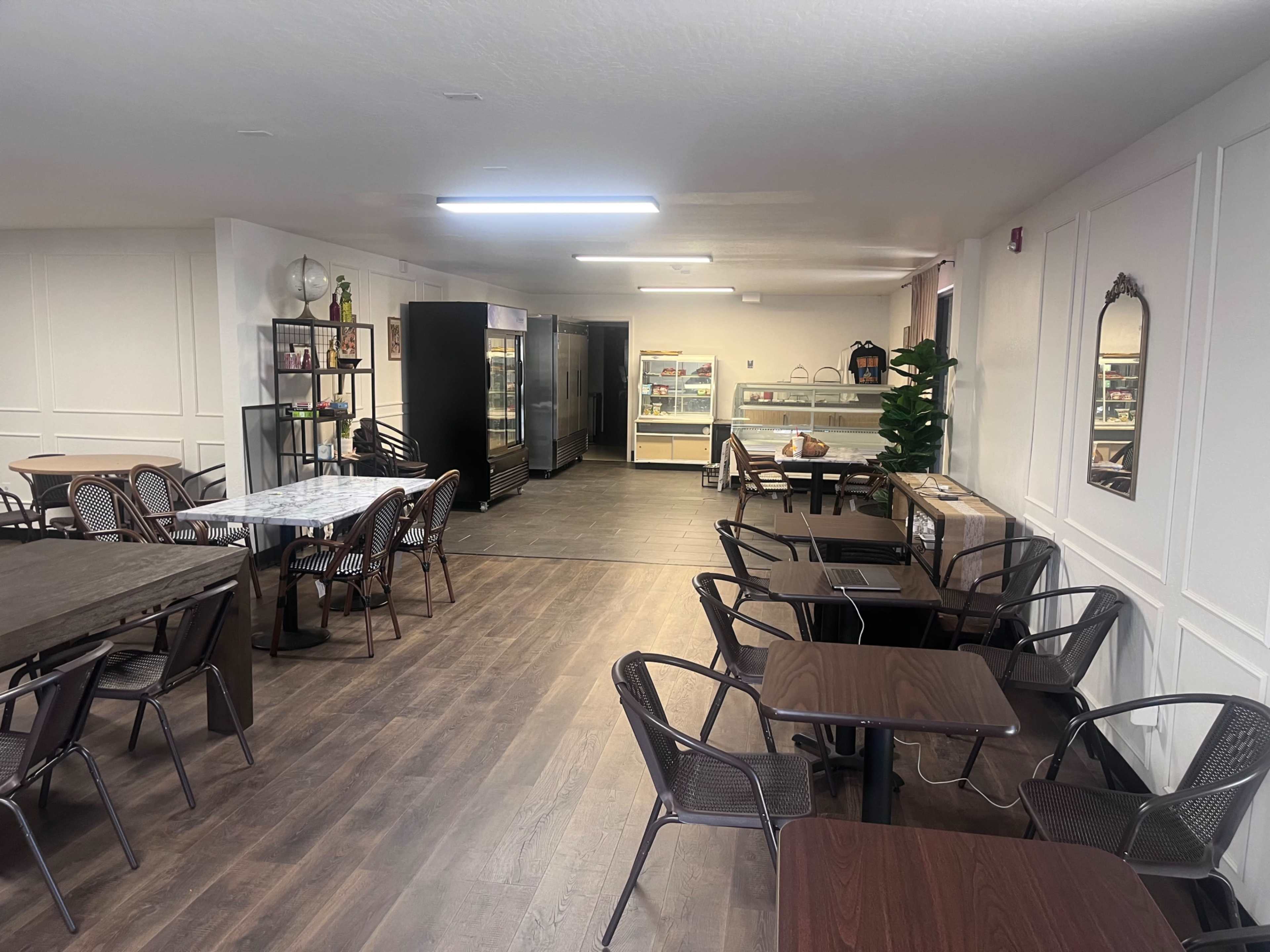 The image shows a spacious café interior with wooden flooring, dining tables, and a display area featuring refrigerated shelves.