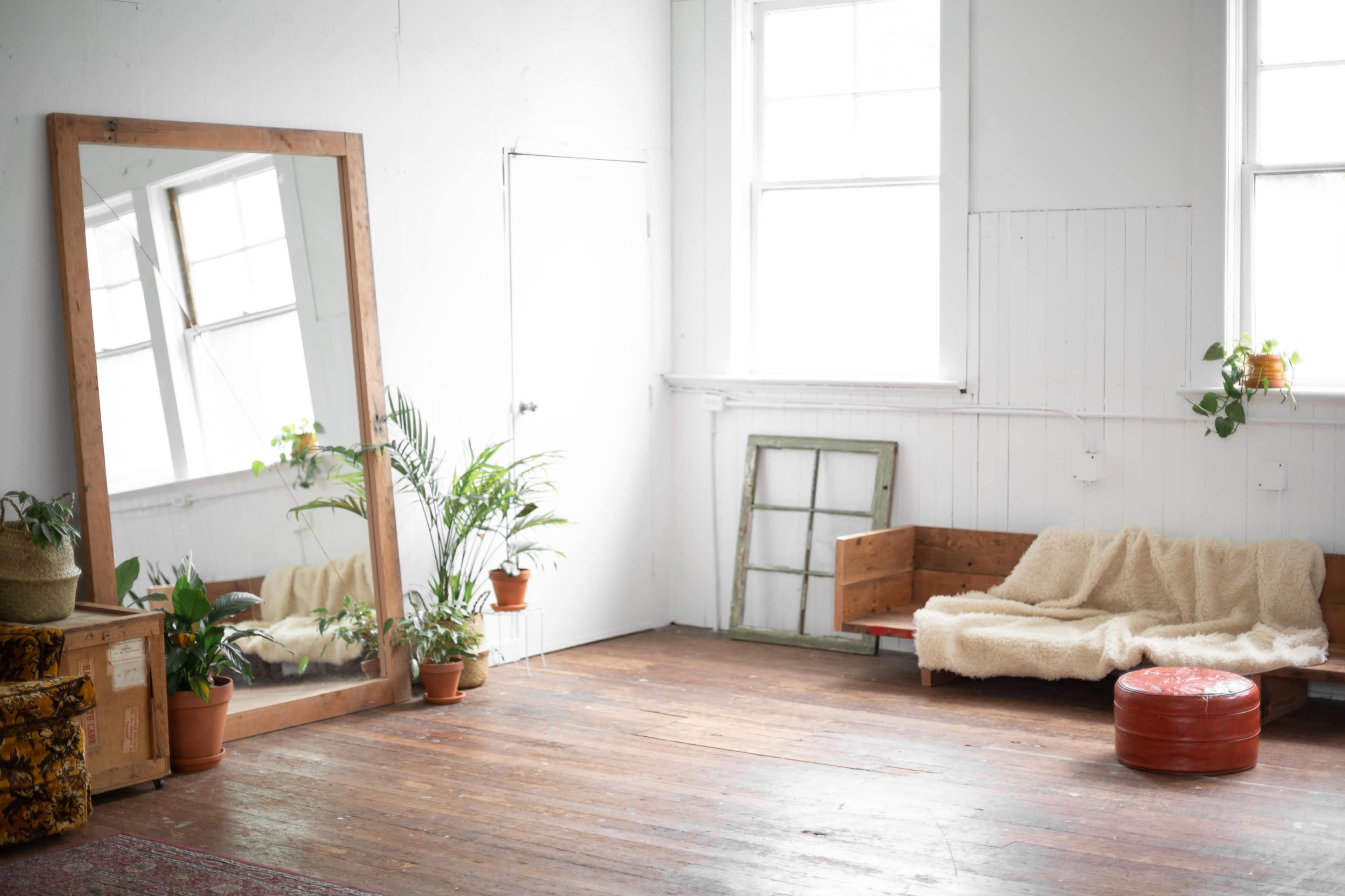 A spacious room features a large mirror, a wooden sofa with a blanket, a round orange ottoman, and several potted plants.