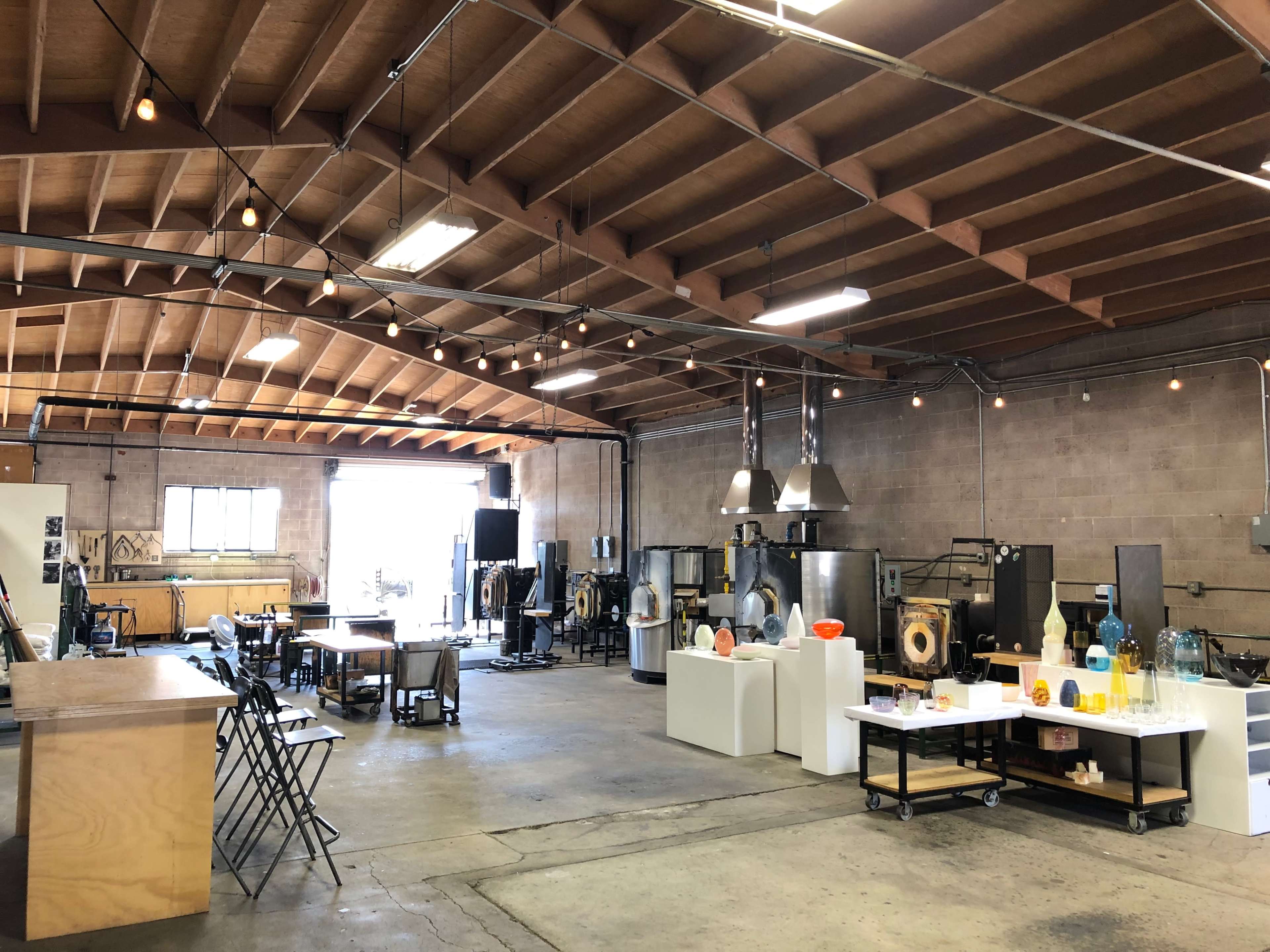 The image shows a spacious art studio with industrial equipment, tables, and display stands for glassblowing or similar craft activities.