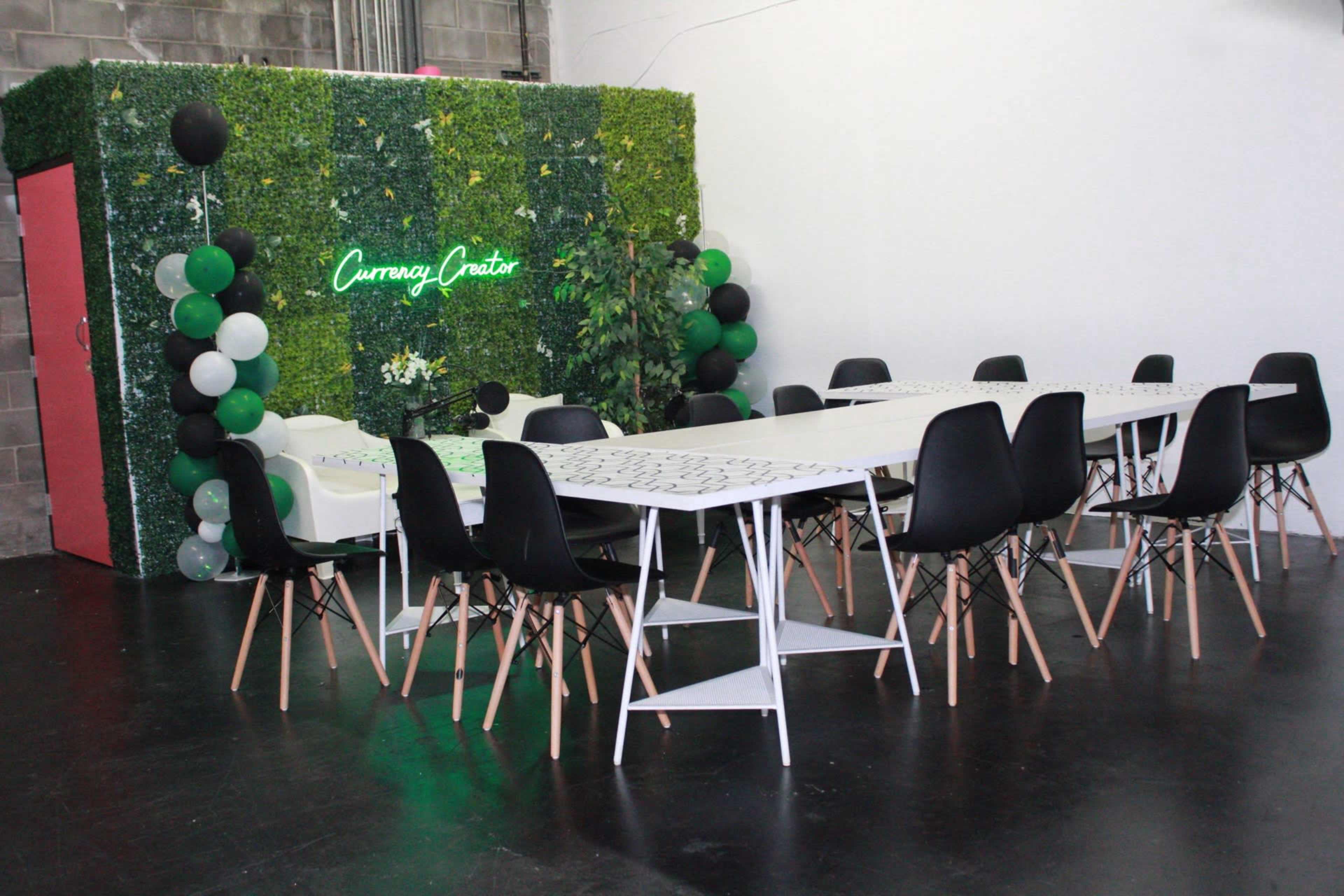 A large, well-lit meeting space features a long white table surrounded by black chairs, with a green decorative wall and a neon sign in the background.