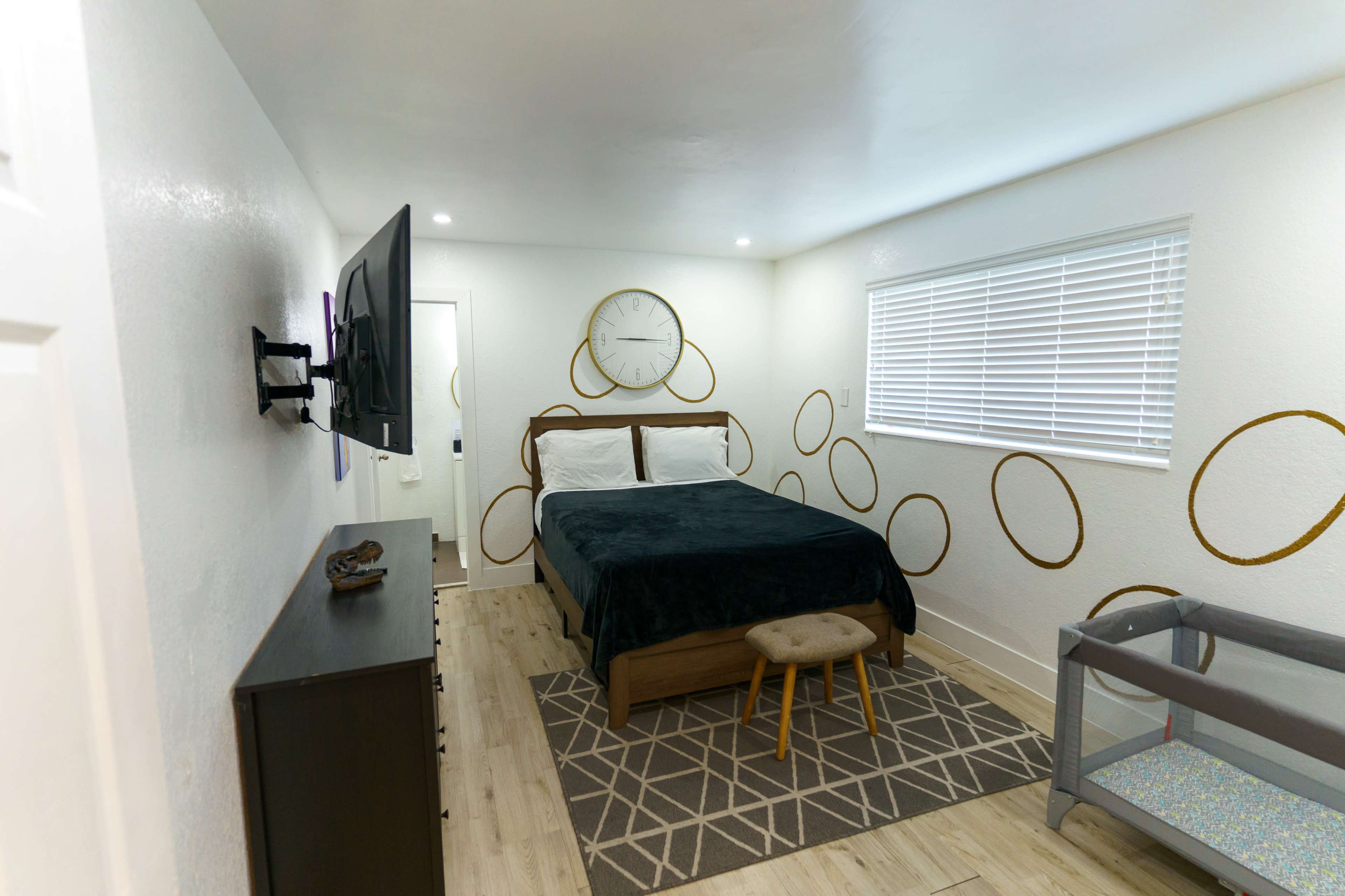 A neatly arranged bedroom featuring a bed with dark bedding, a wall-mounted television, a modern clock, and a small changing table.