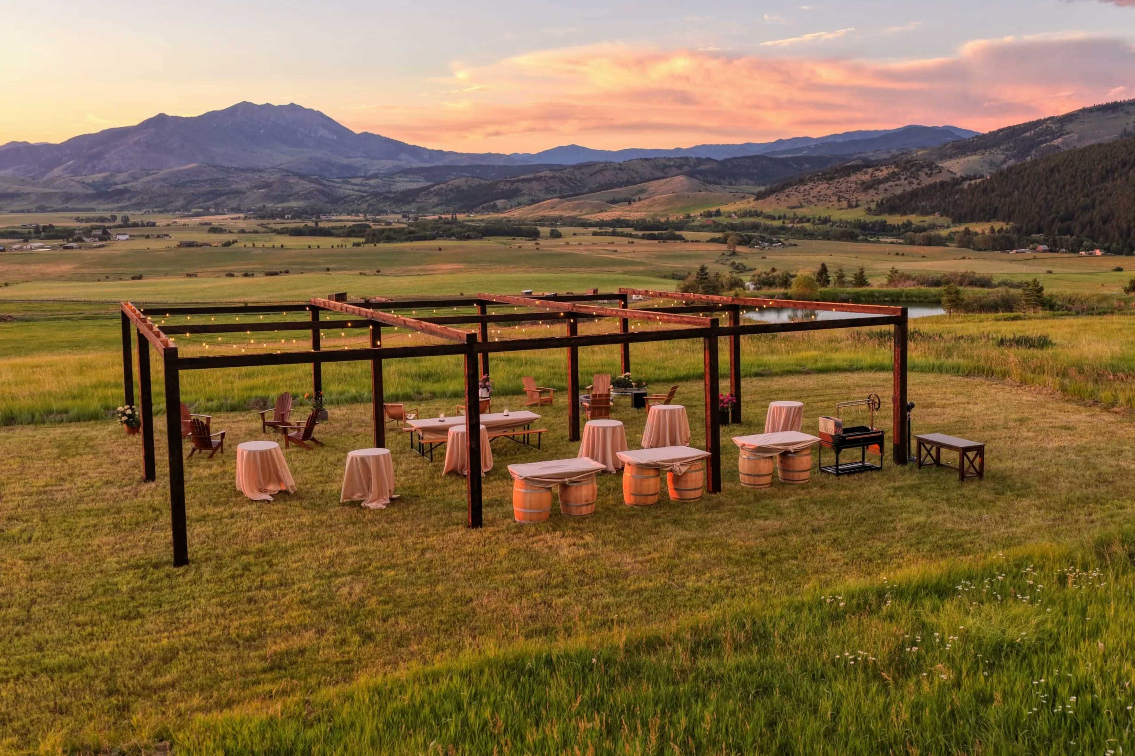 An outdoor event space with several tables and chairs is set up on a grassy area, framed by mountains and a sunset sky.