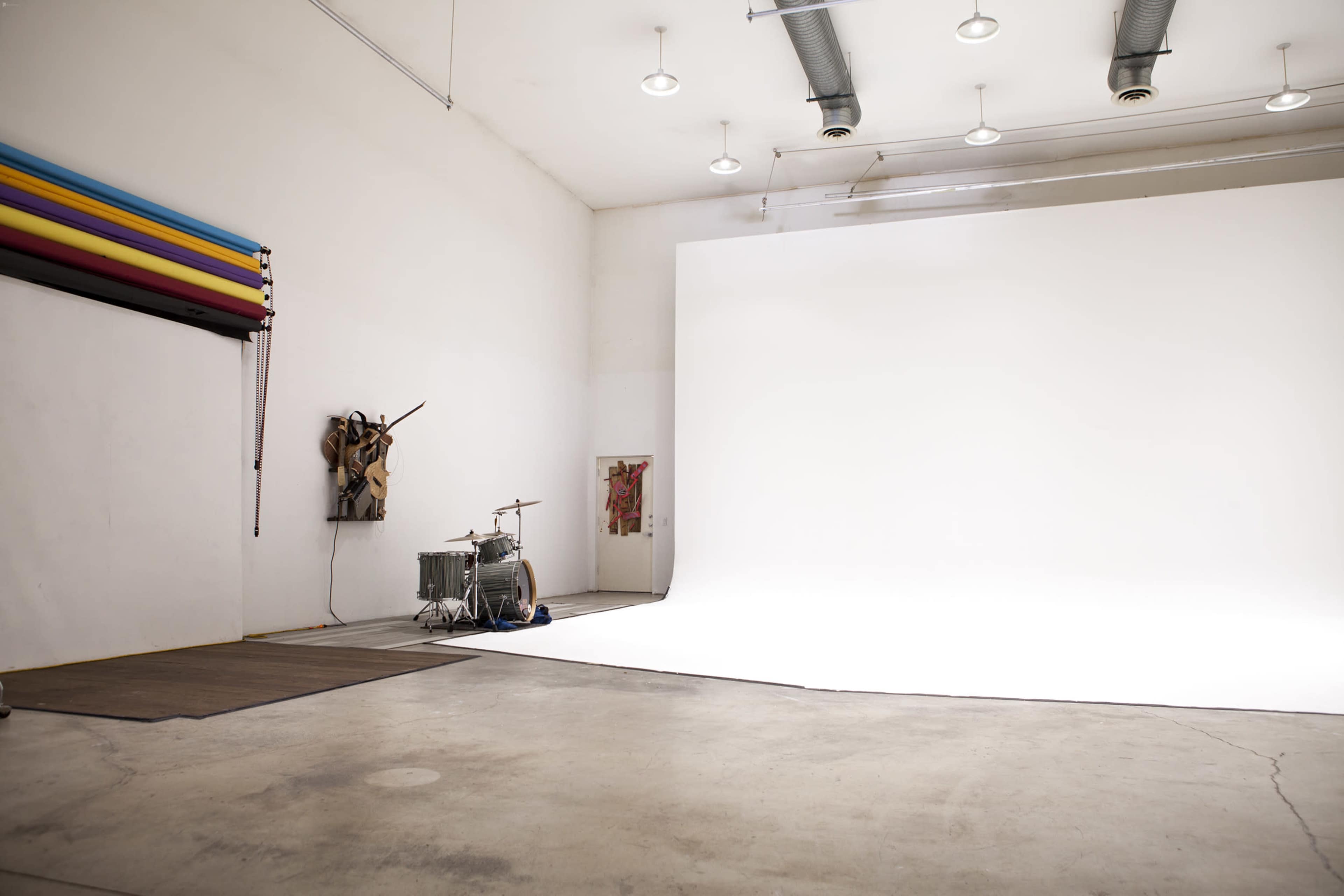 A spacious photography studio features a large white backdrop, a wooden floor, and a drum set positioned in the corner.