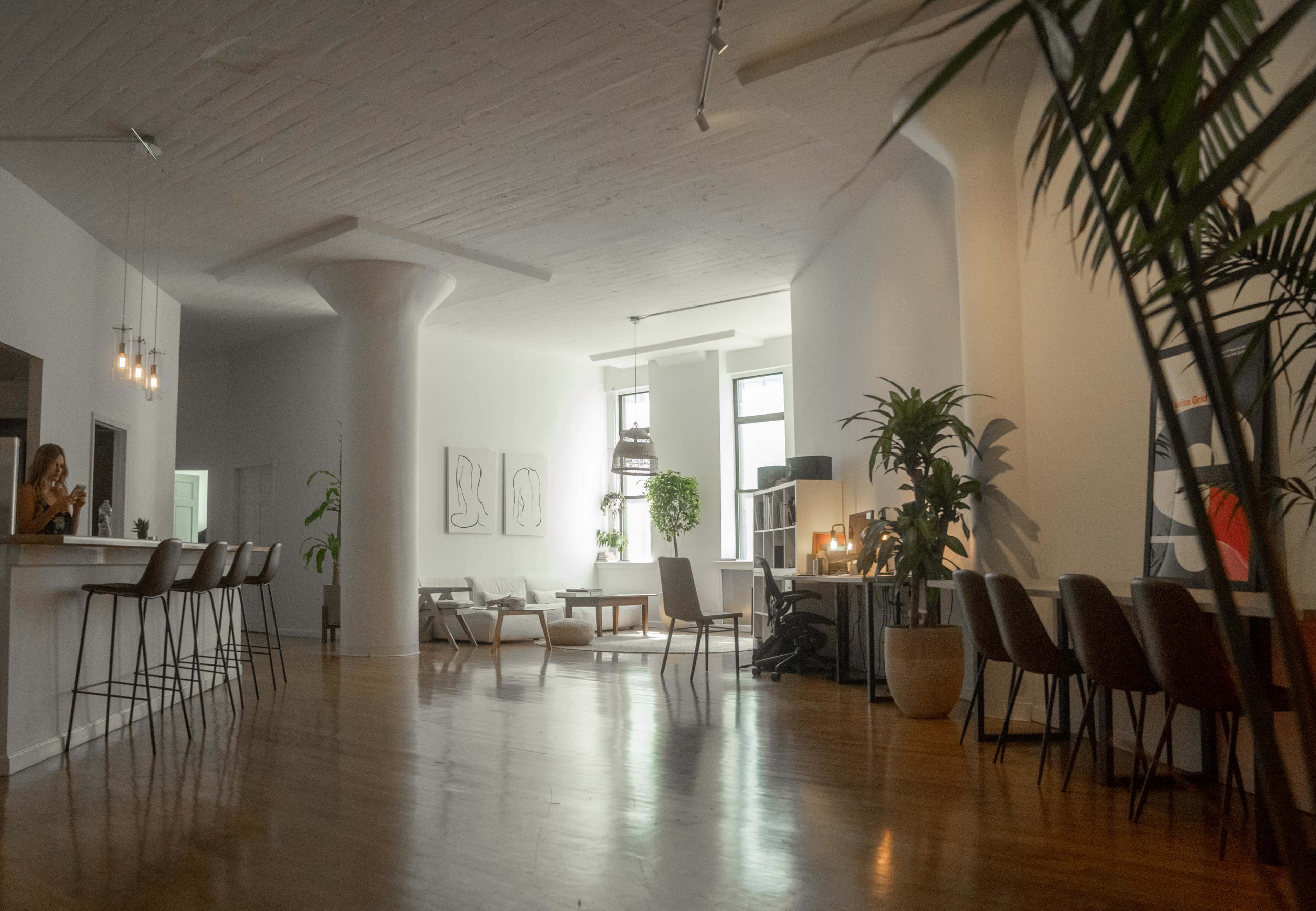 A spacious, minimalist interior with tall ceilings, large windows, and various plants, featuring a bar counter, seating area, and artwork on the walls.