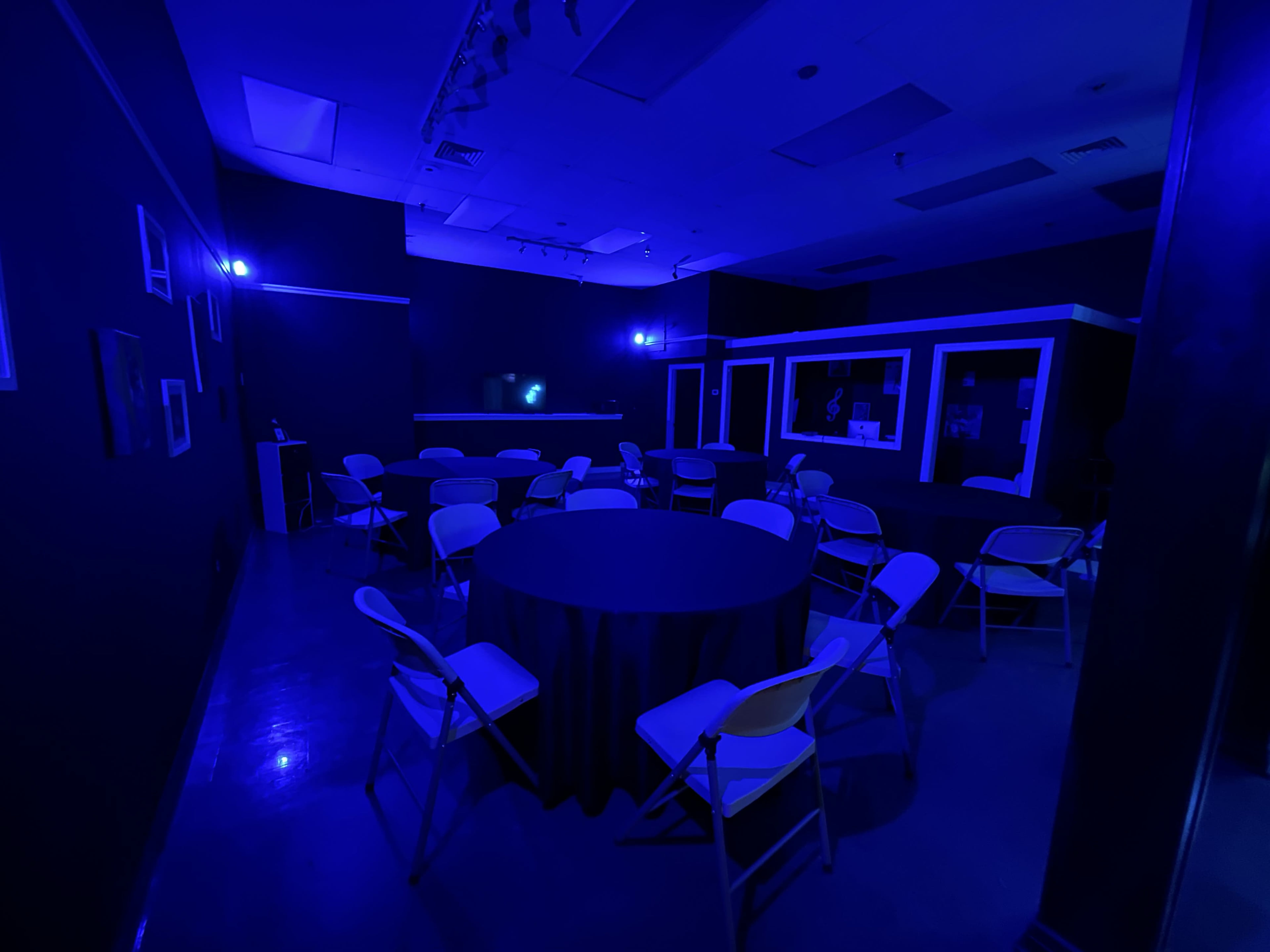 A dimly lit room with blue lighting, featuring a round table surrounded by folding chairs and a bar area in the background.