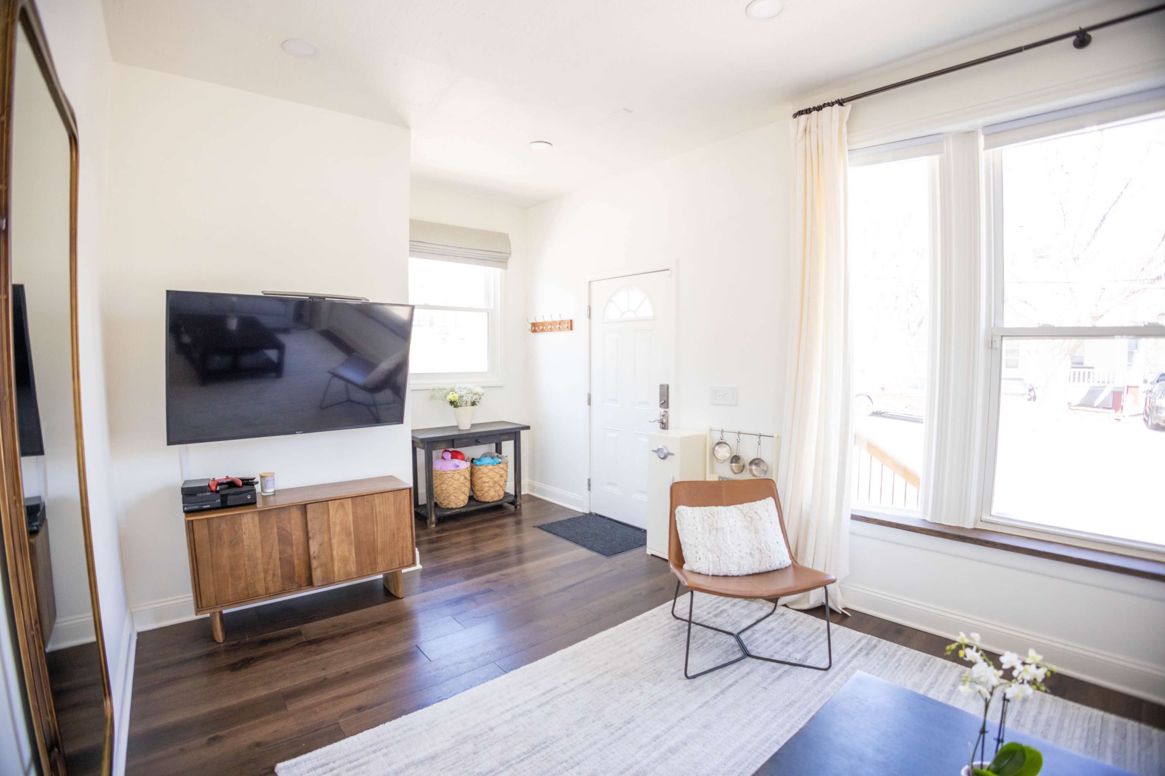 The image shows a modern living room featuring a television mounted on the wall, a stylish wooden cabinet, a door leading outside, and a cozy armchair on a light-colored rug.