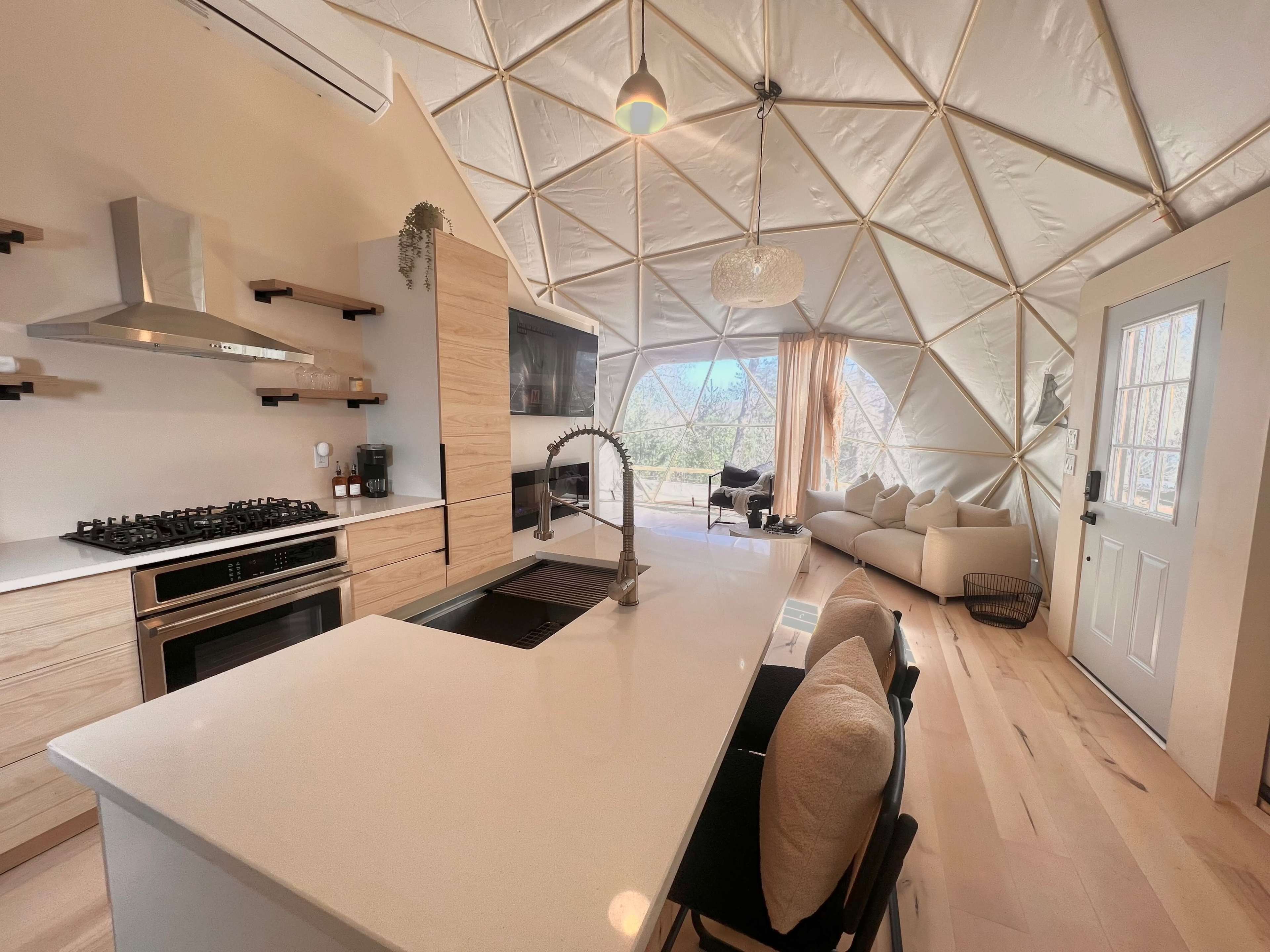 A modern kitchen and living area within a geodesic dome featuring light-colored wood cabinetry, a large island with a sink, and a cozy seating area with a view through large windows.
