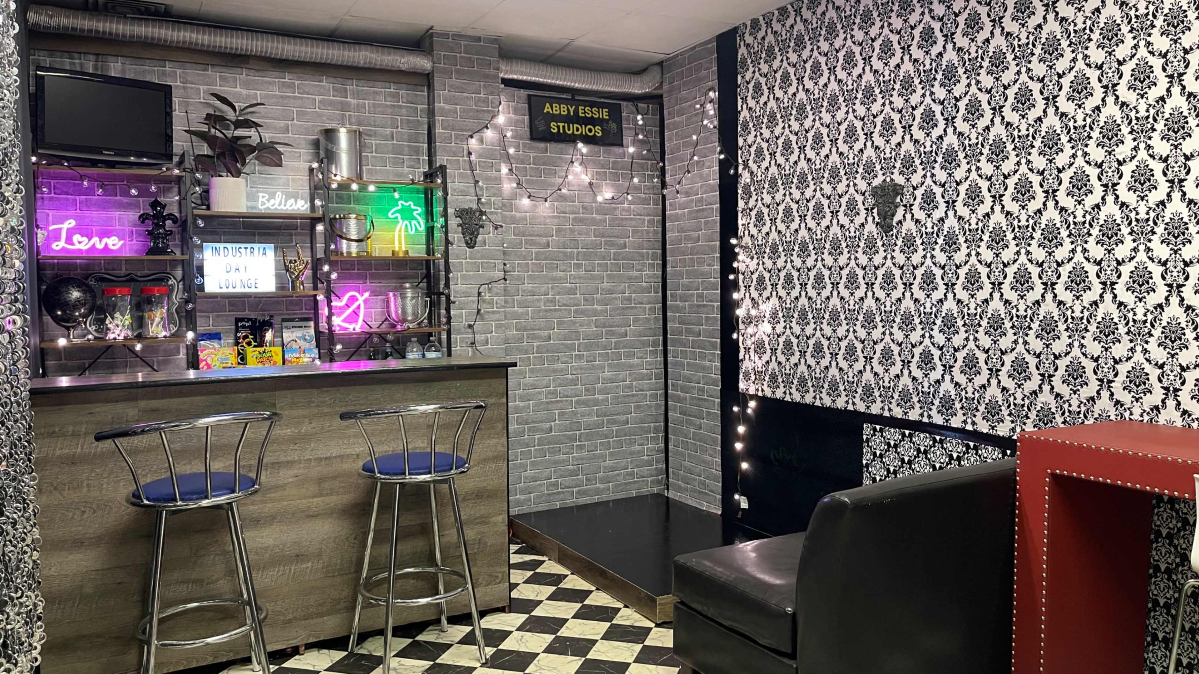 The image shows a small studio space with a bar area featuring neon signs, a checkerboard floor, and decorative wallpaper.