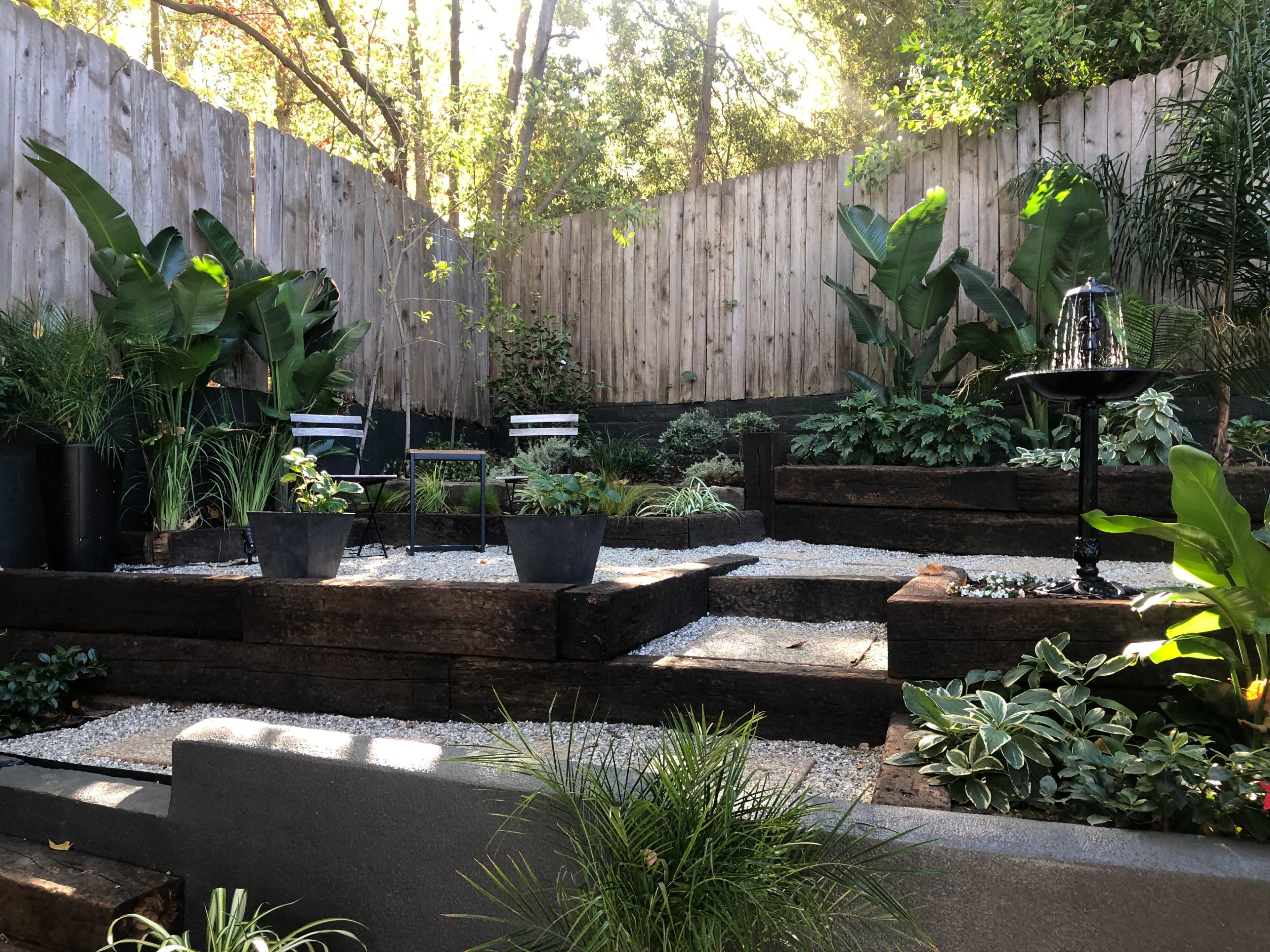 A landscaped garden with multiple levels, featuring potted plants, gravel paths, and wooden planters surrounded by greenery and a wooden fence.