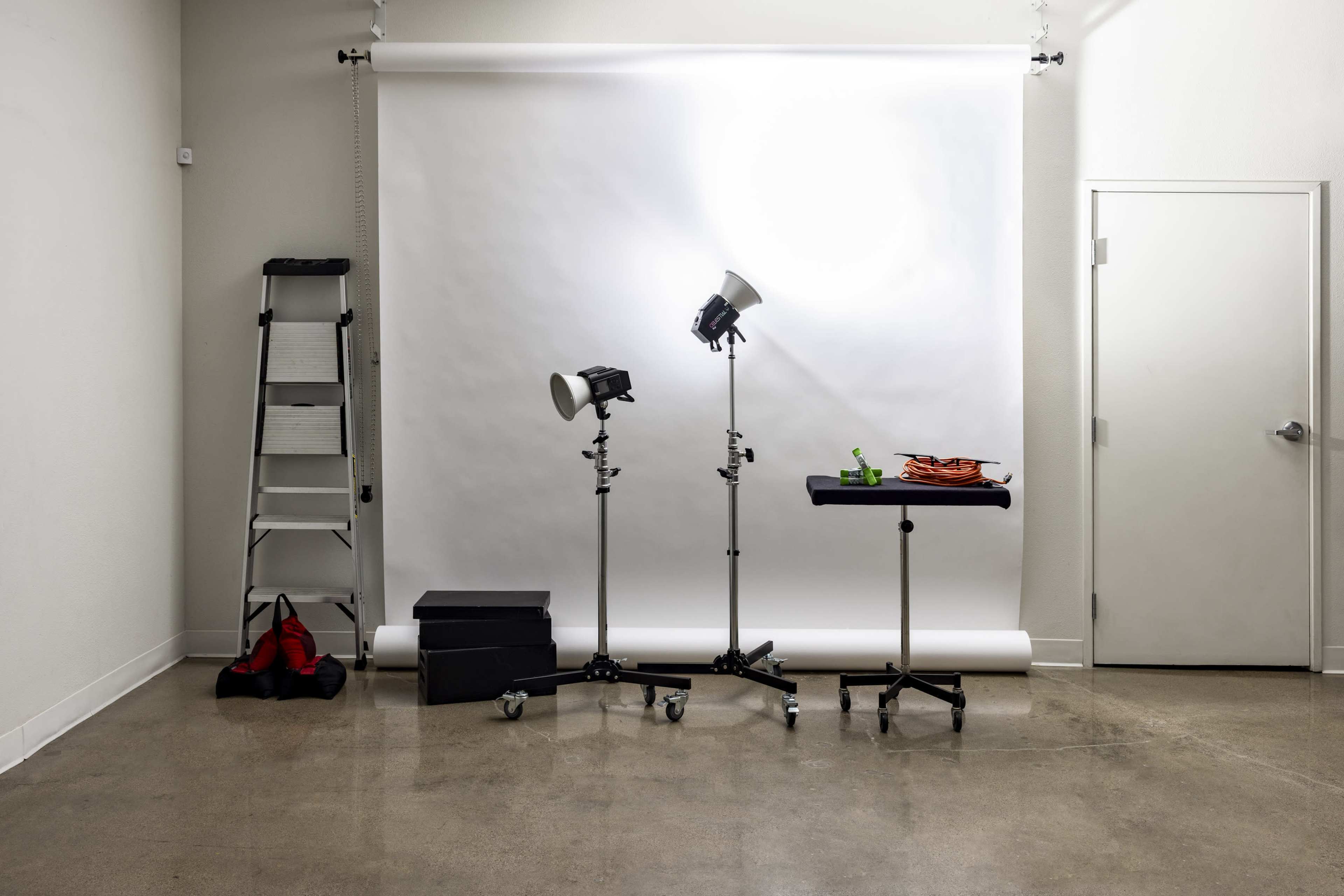 The image shows a photography studio setting, featuring a white backdrop, lighting equipment, a black table with props, a step ladder, and several storage boxes.