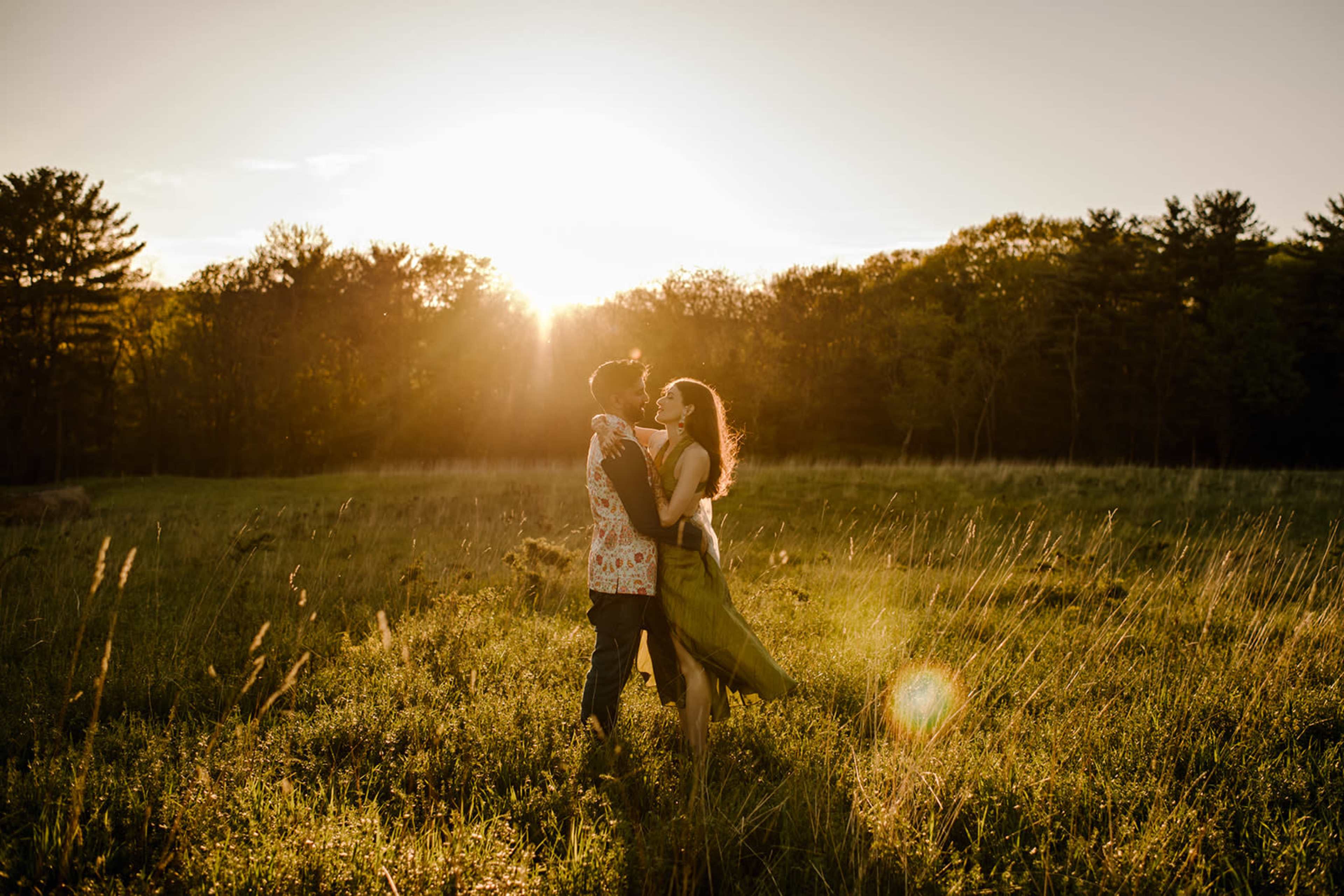 A couple embraces in a grassy field at sunset, with trees and a warm glow in the background.