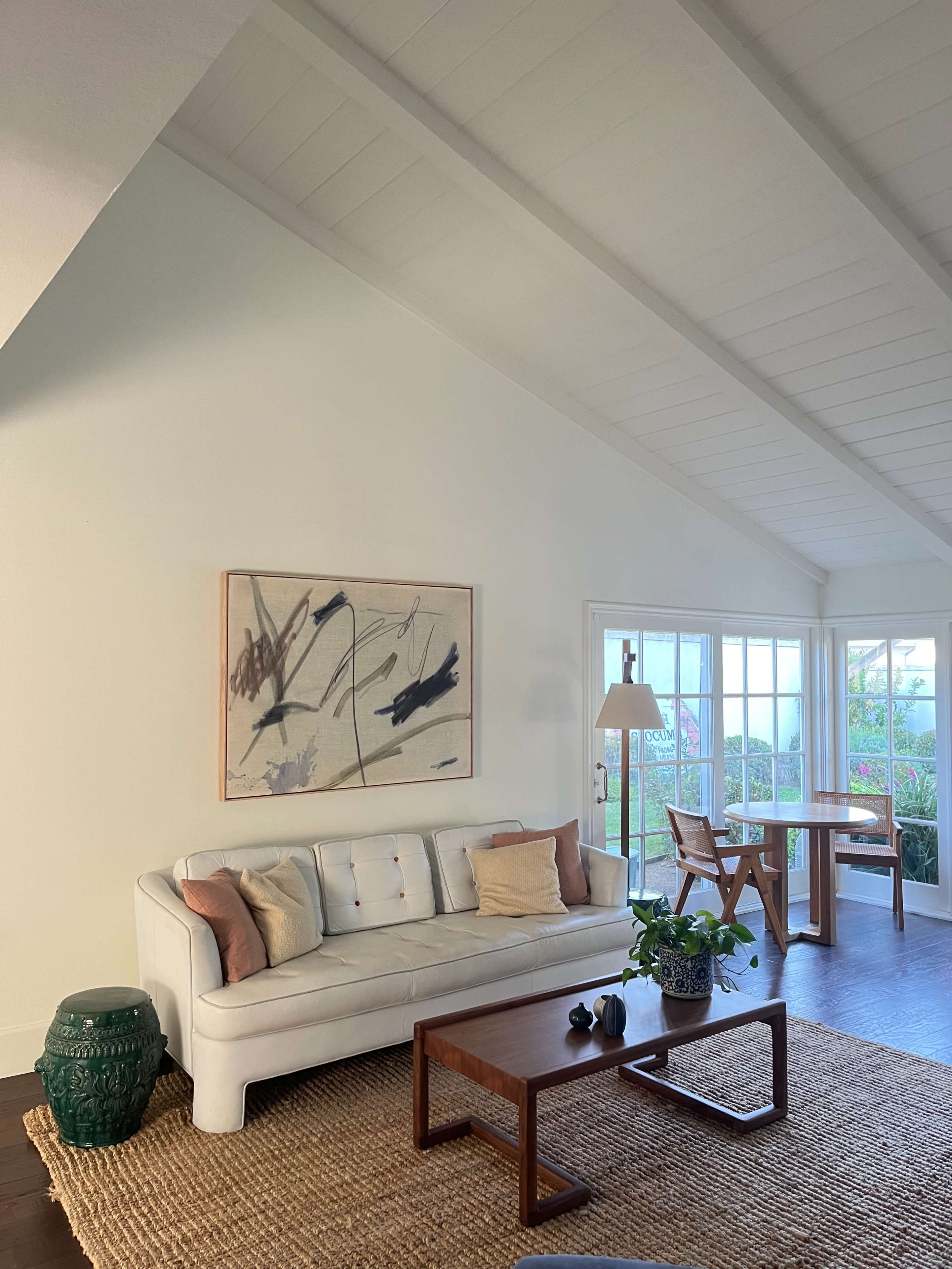 The image shows a bright living room with a white sofa, a wooden coffee table, and a large window providing a view of the outdoors.