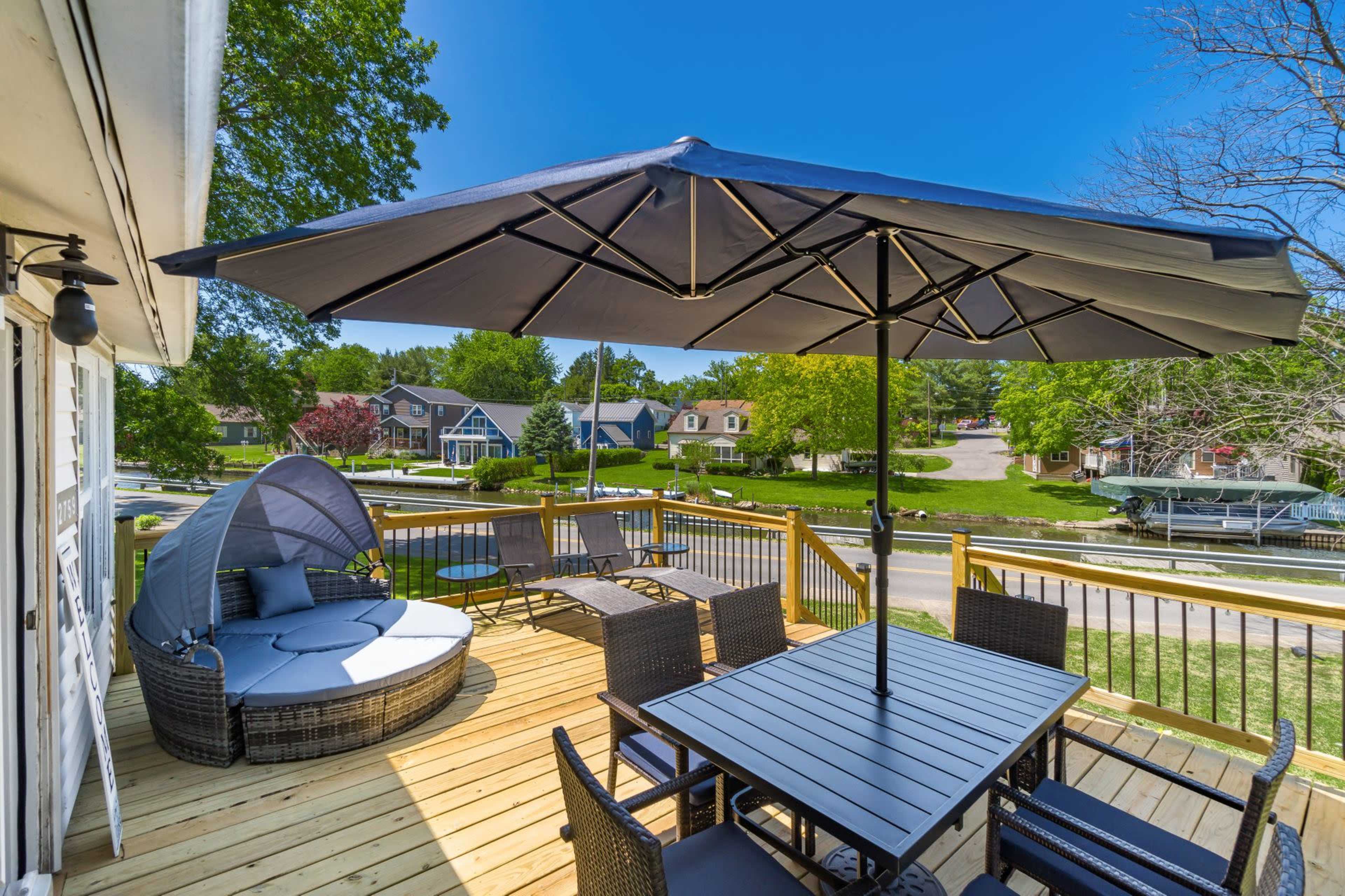A wooden deck features a circular seating area with a large umbrella, a dining table set, and a view of nearby houses and greenery.