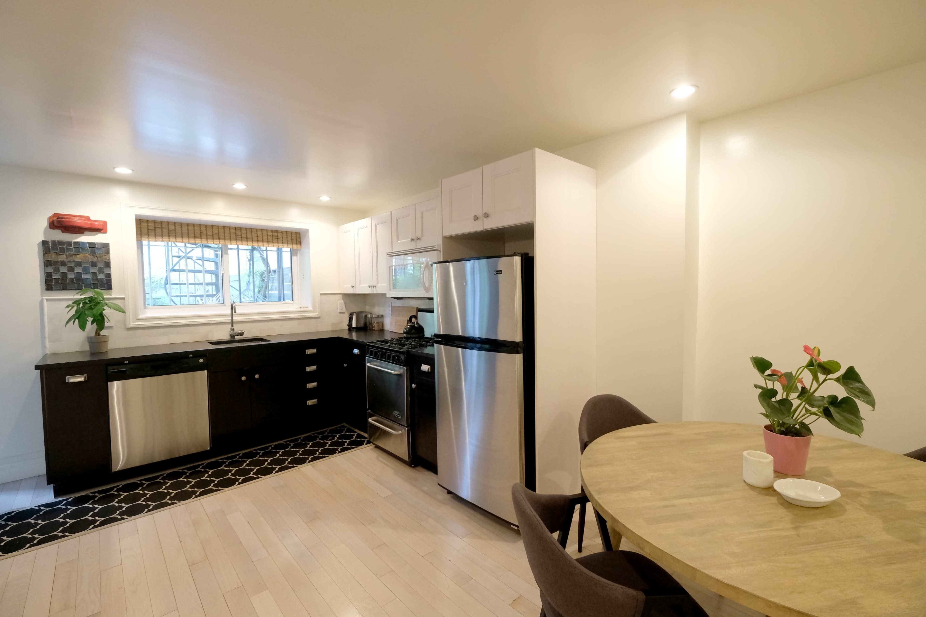 The image shows a modern kitchen with white cabinetry, stainless steel appliances, and a small dining area featuring a round table and a potted plant.