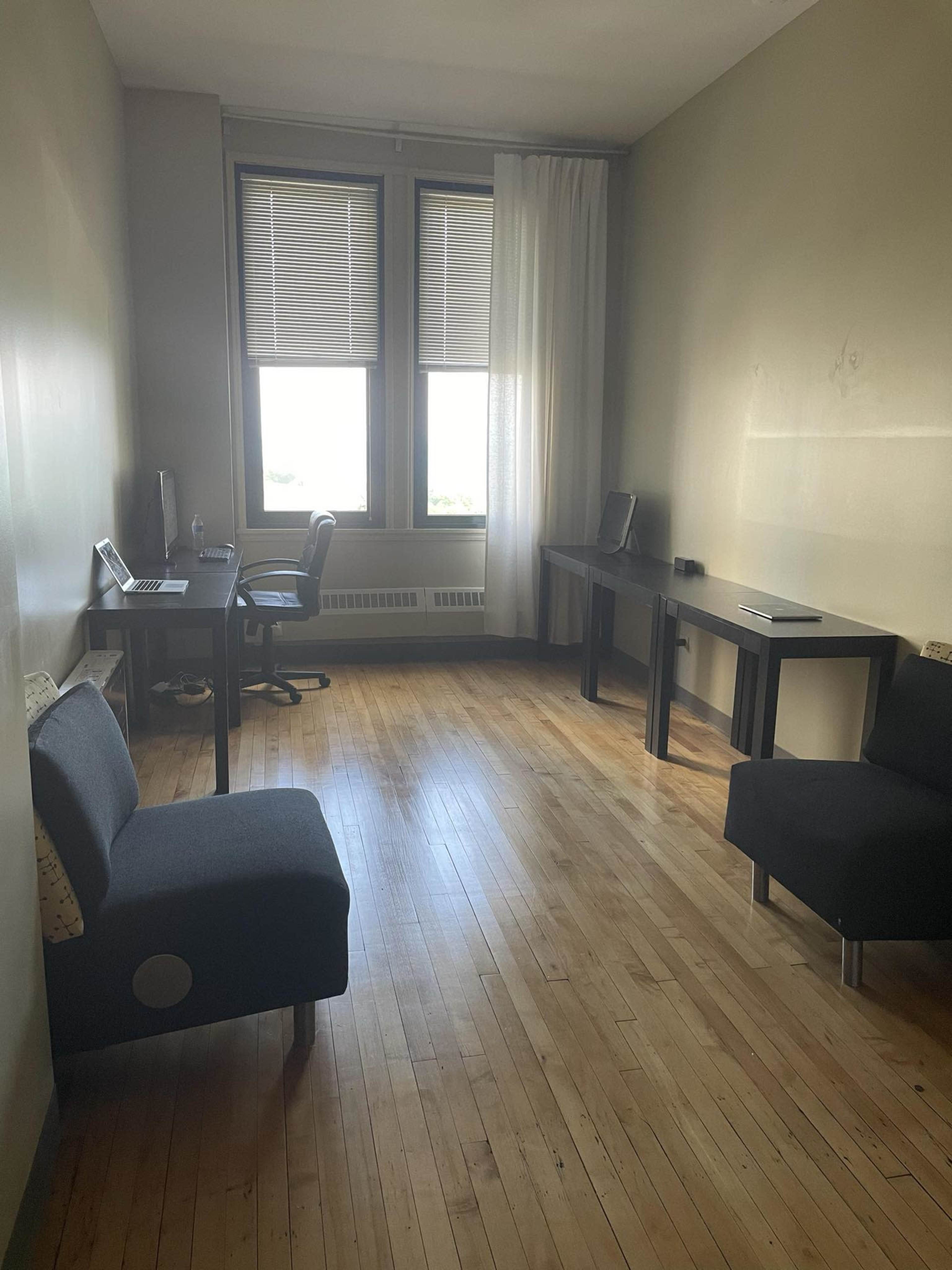 The image shows a sparsely furnished room with wooden floors, featuring a desk and chair, two small sofas, and large windows allowing natural light.