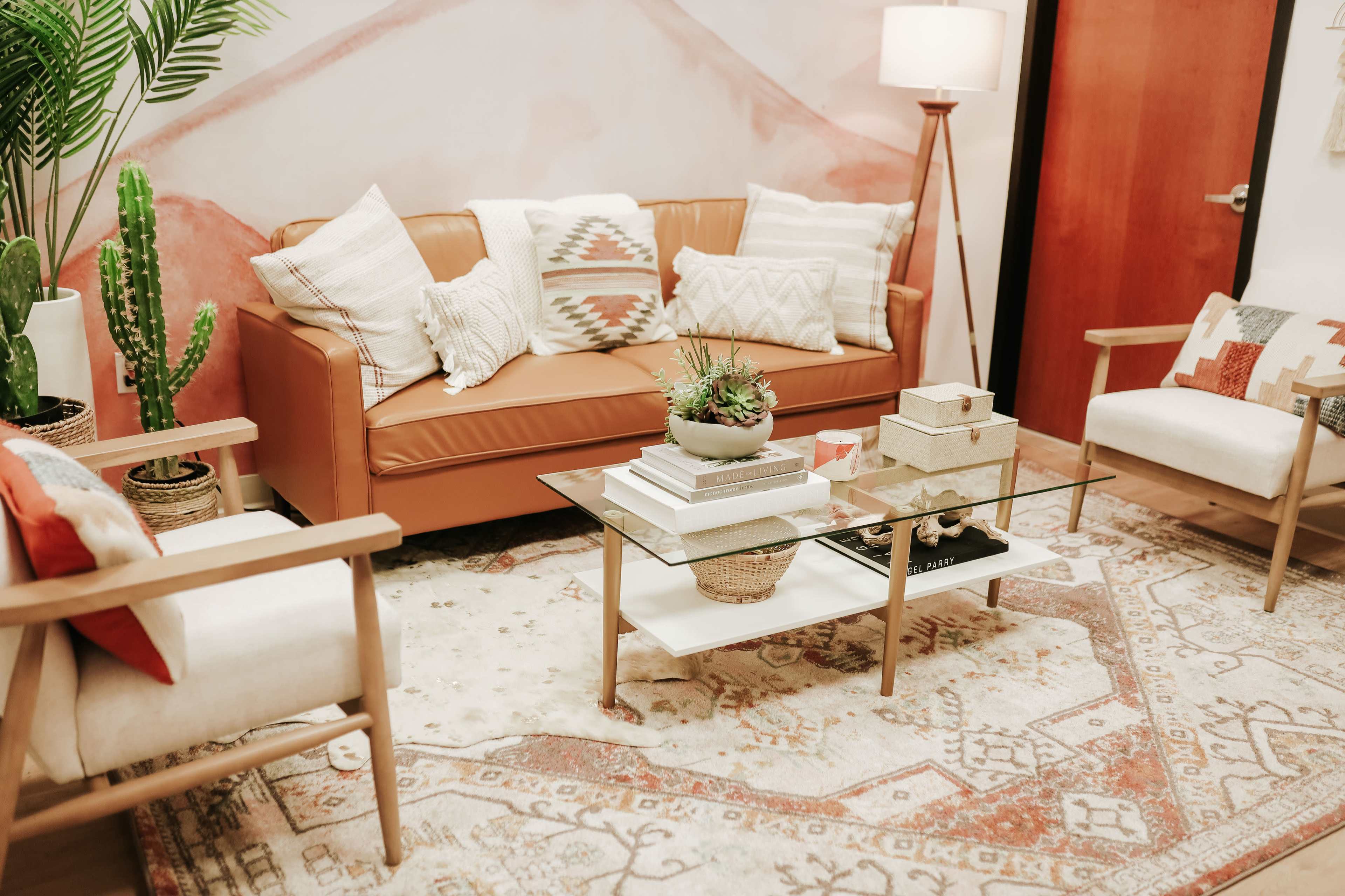 A cozy living room setting with a brown leather sofa, a glass coffee table, and various decorative elements, including plants and patterned cushions, arranged on a patterned rug.