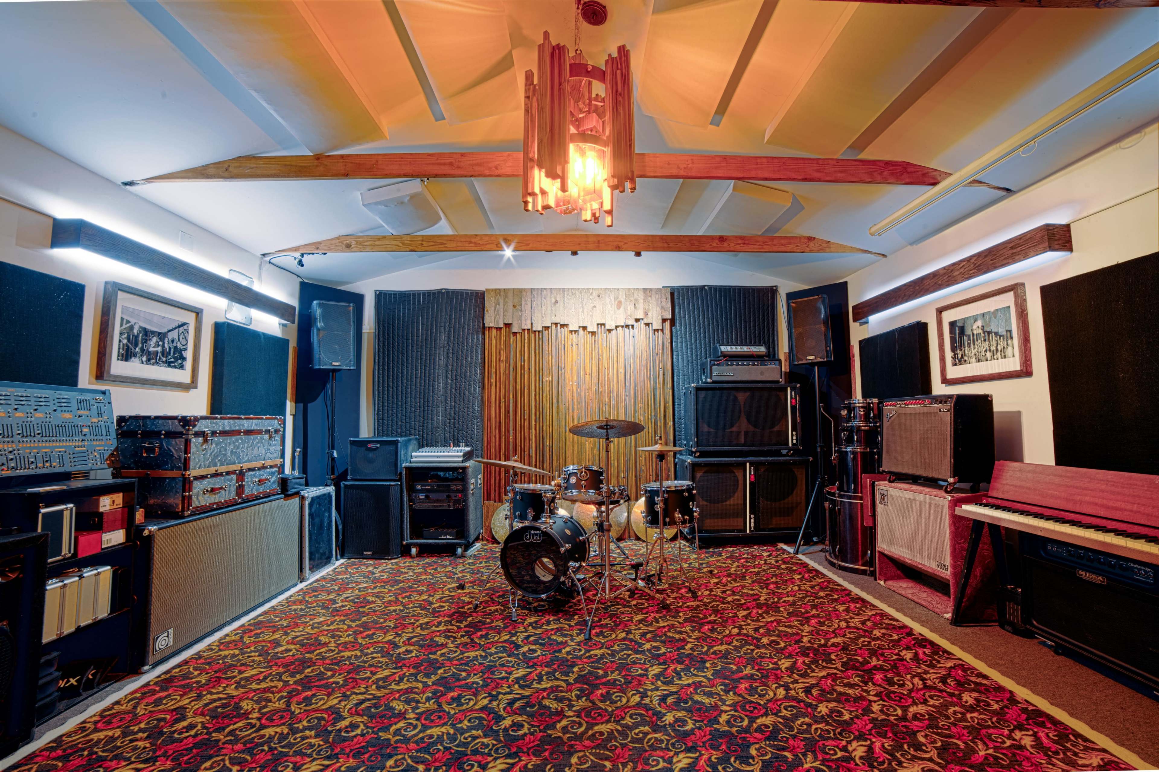 The image shows a music studio with a drum set in the center, surrounded by various amplifiers and speakers against patterned carpet and wood-paneled walls.