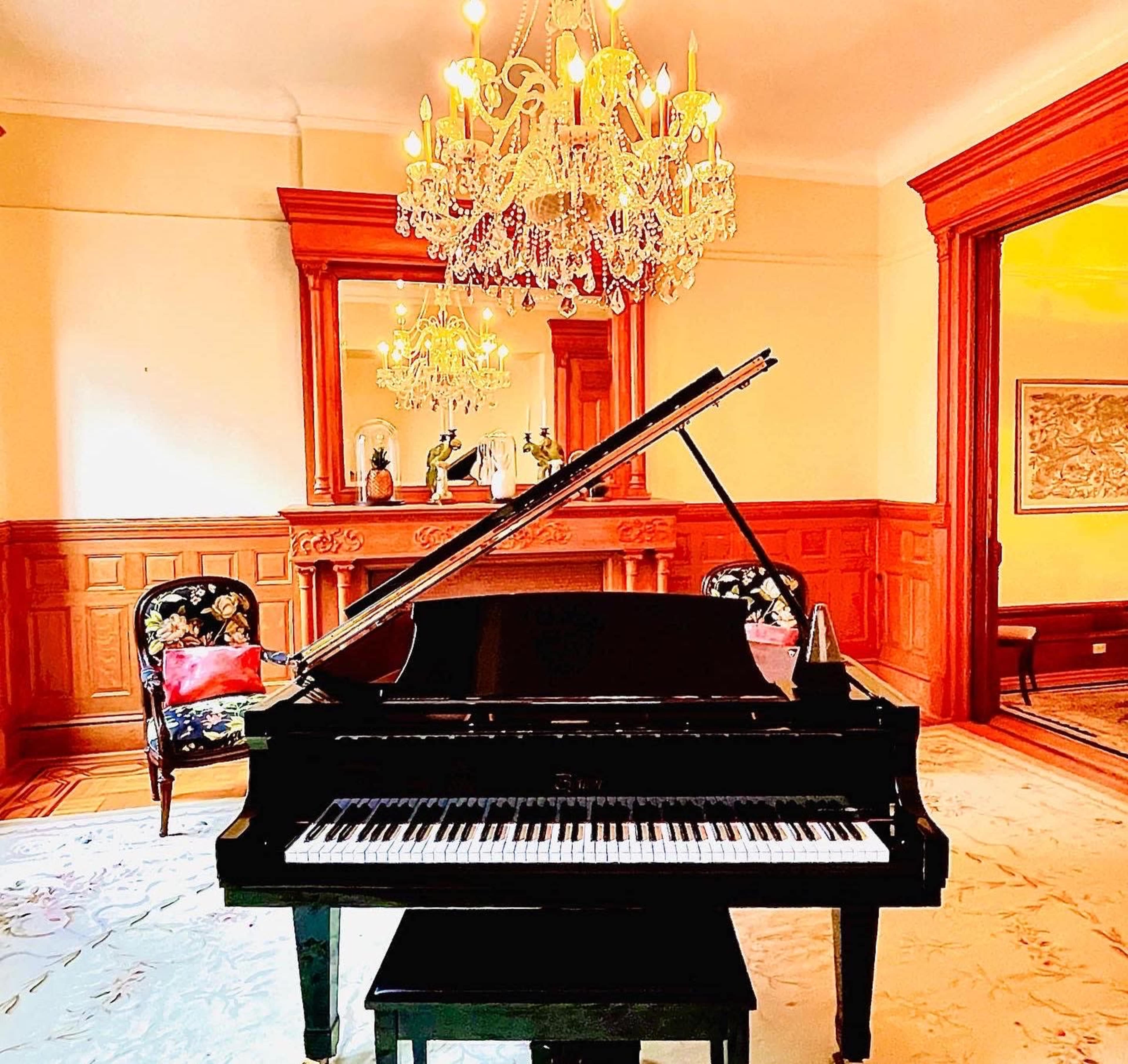 A grand piano is positioned in front of a large chandelier and an ornate mirror in a room with wooden paneling.