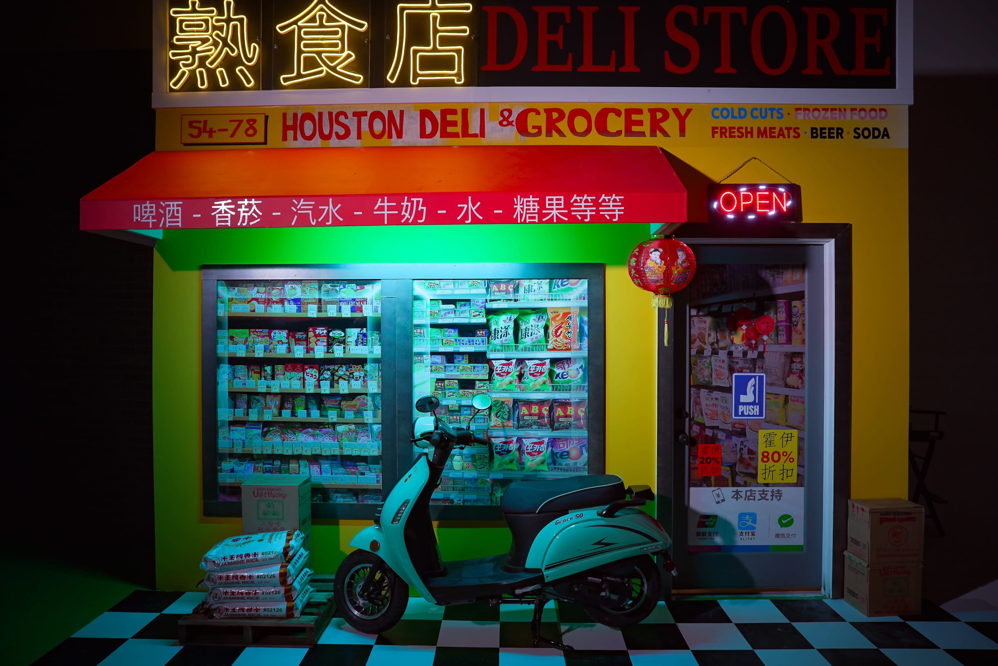 A brightly lit deli store displays a variety of groceries and cold cuts, with a scooter parked in front of its entrance.