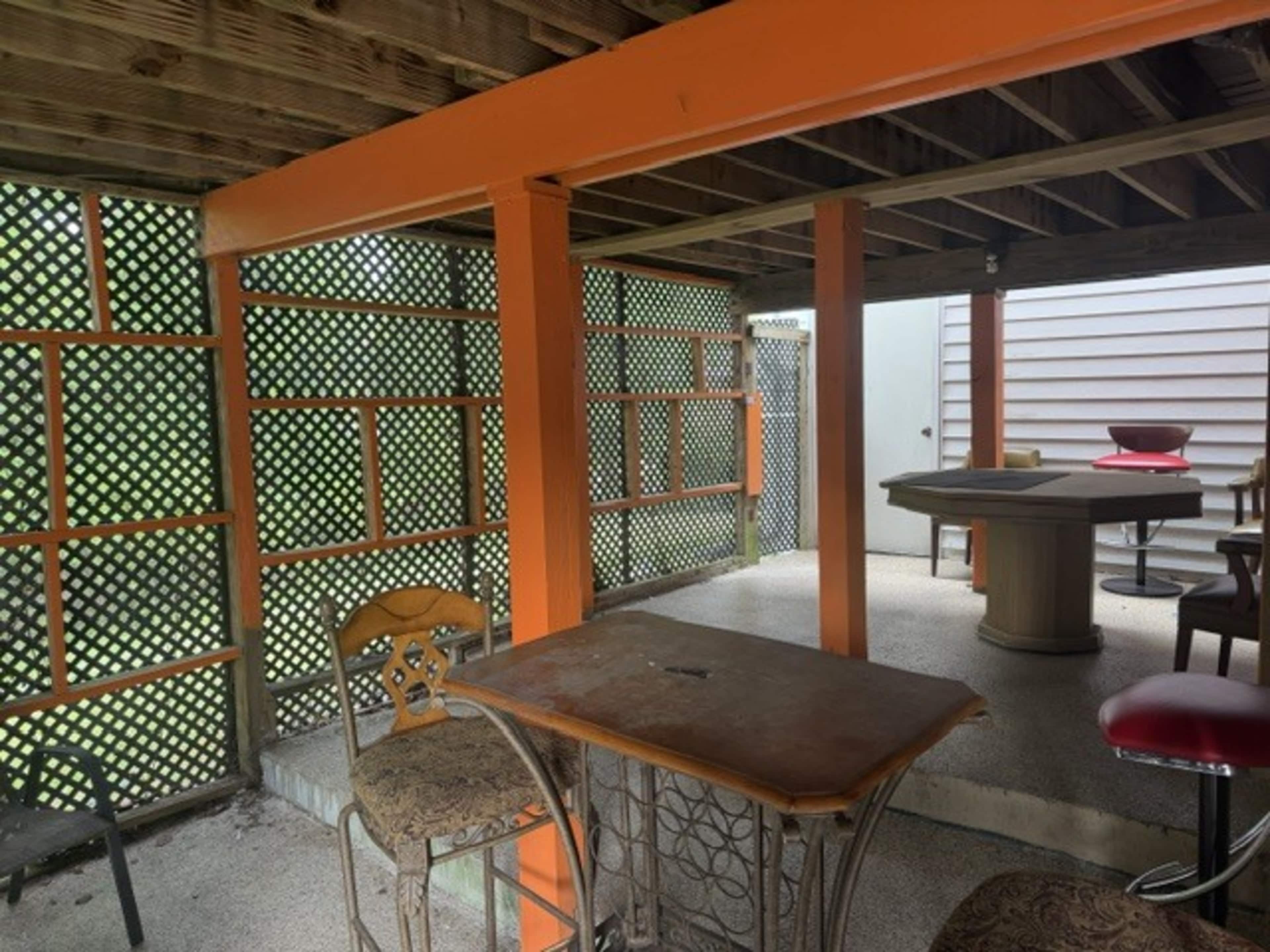 The image shows a partially covered outdoor space with an orange-beamed ceiling, a table with chairs, and a high-top bar area against a lattice backdrop.