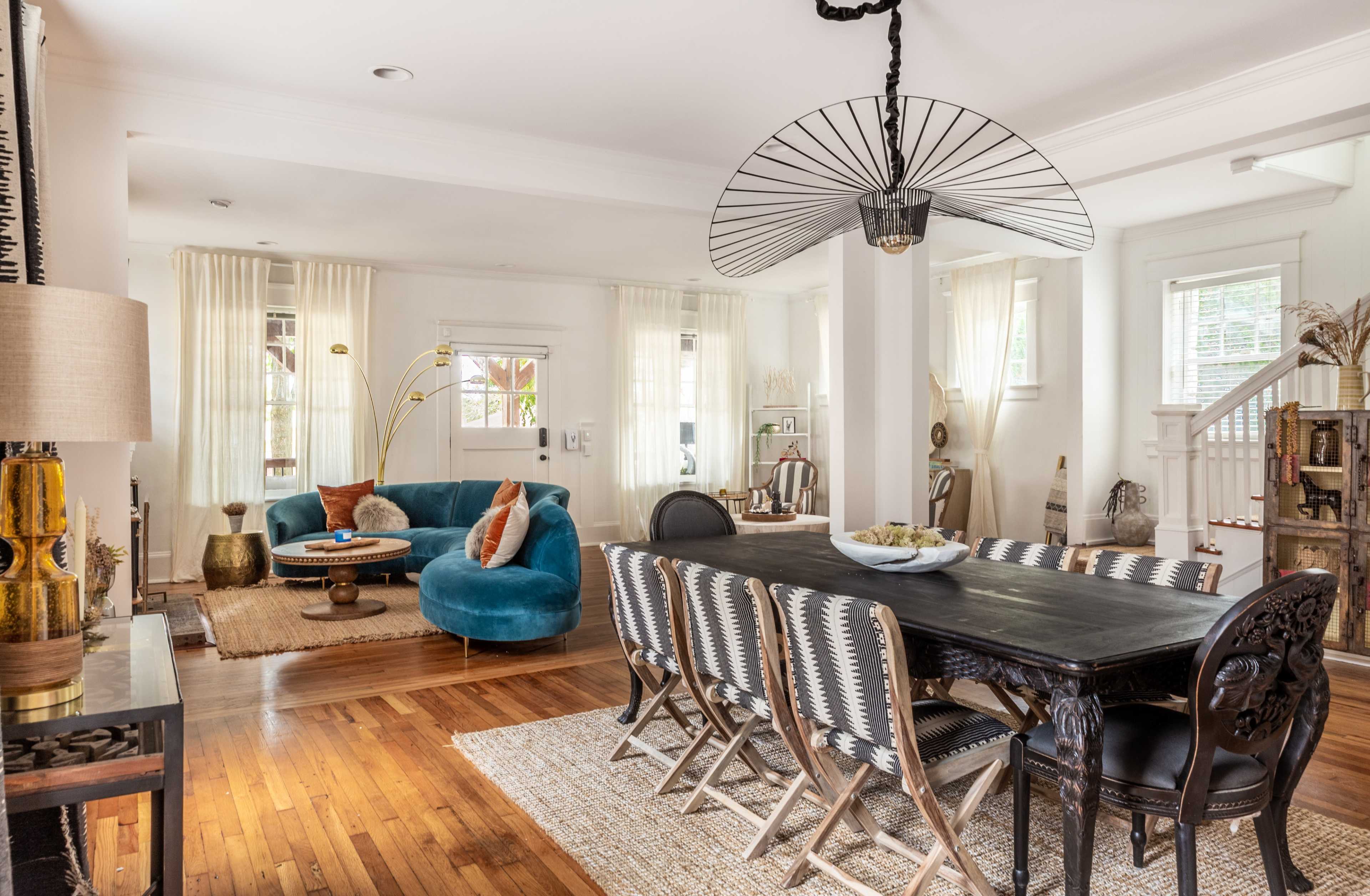 A spacious living and dining area with a turquoise sofa, a long dark wooden dining table, and bright curtains allowing natural light to illuminate the room.