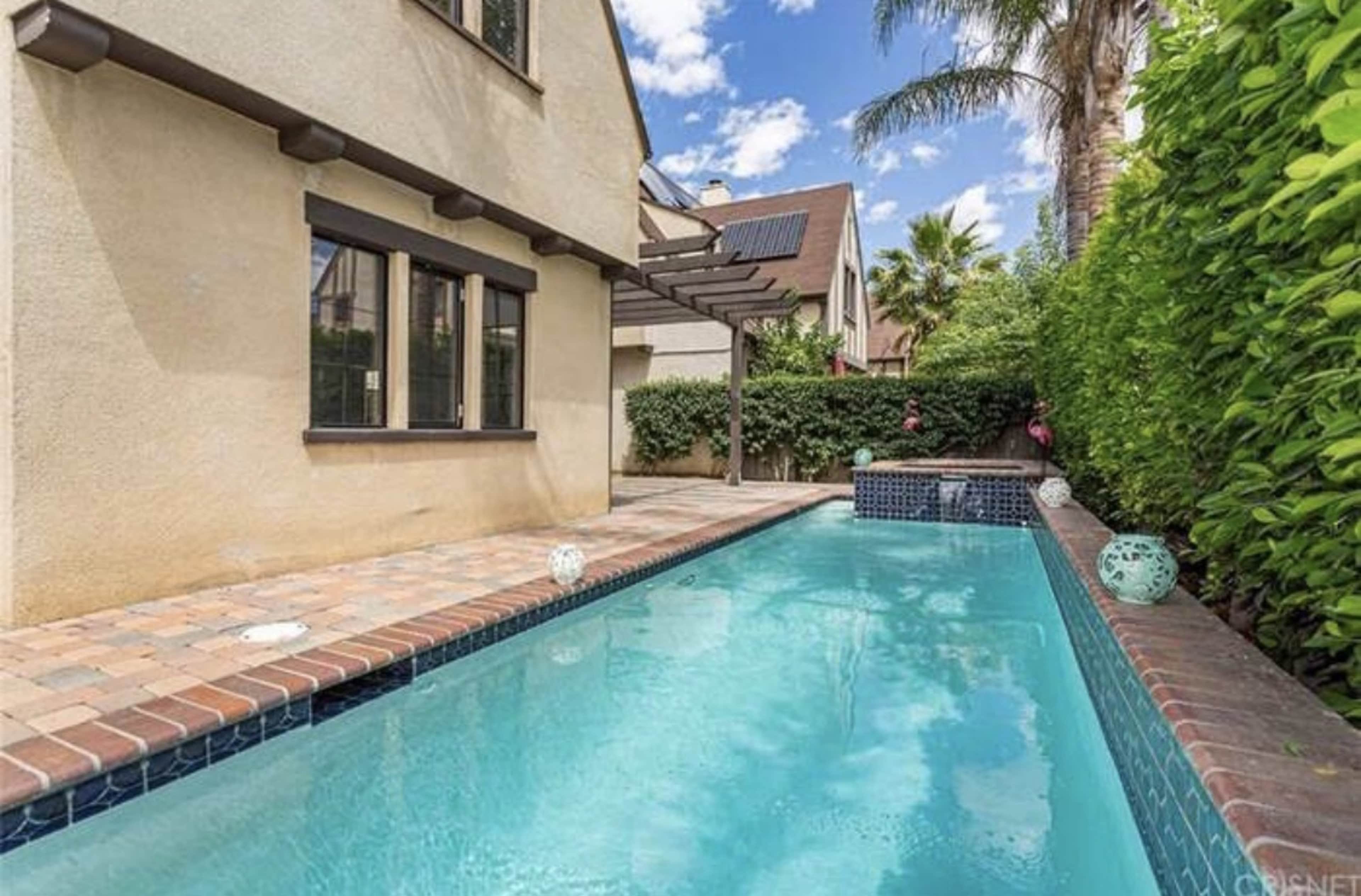 A narrow swimming pool bordered by brick and surrounded by greenery, adjacent to a house with a pergola.