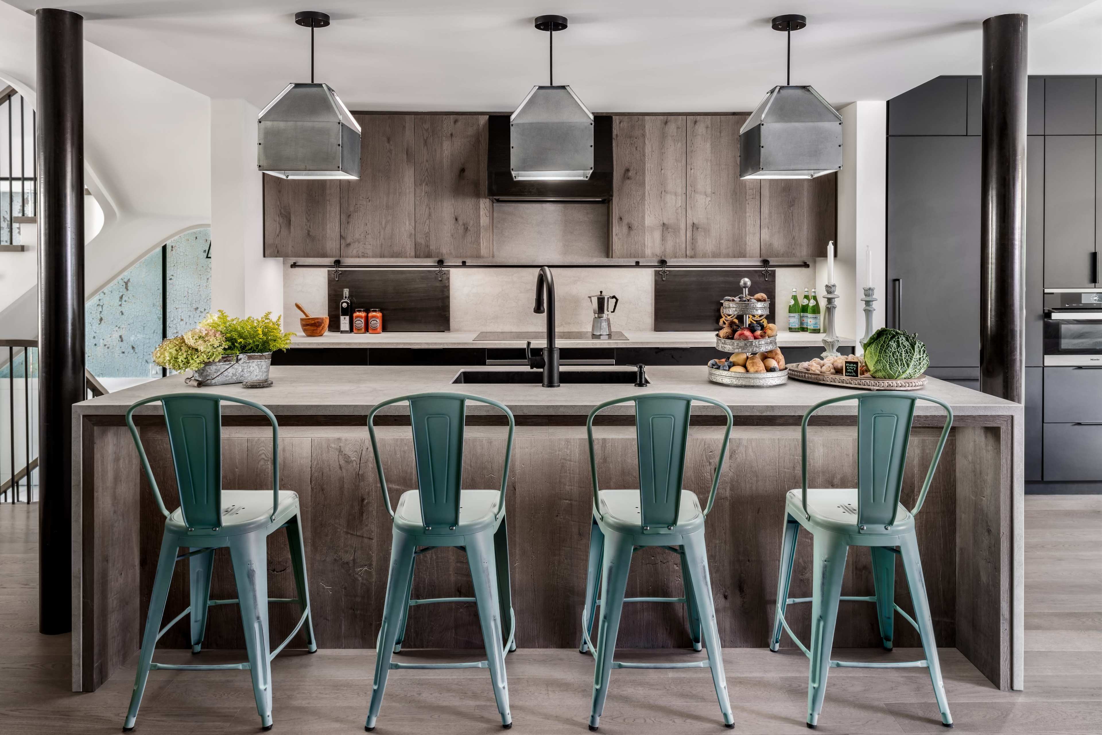 A modern kitchen features a large island with four metal stools, dark wood cabinetry, and pendant lights above.