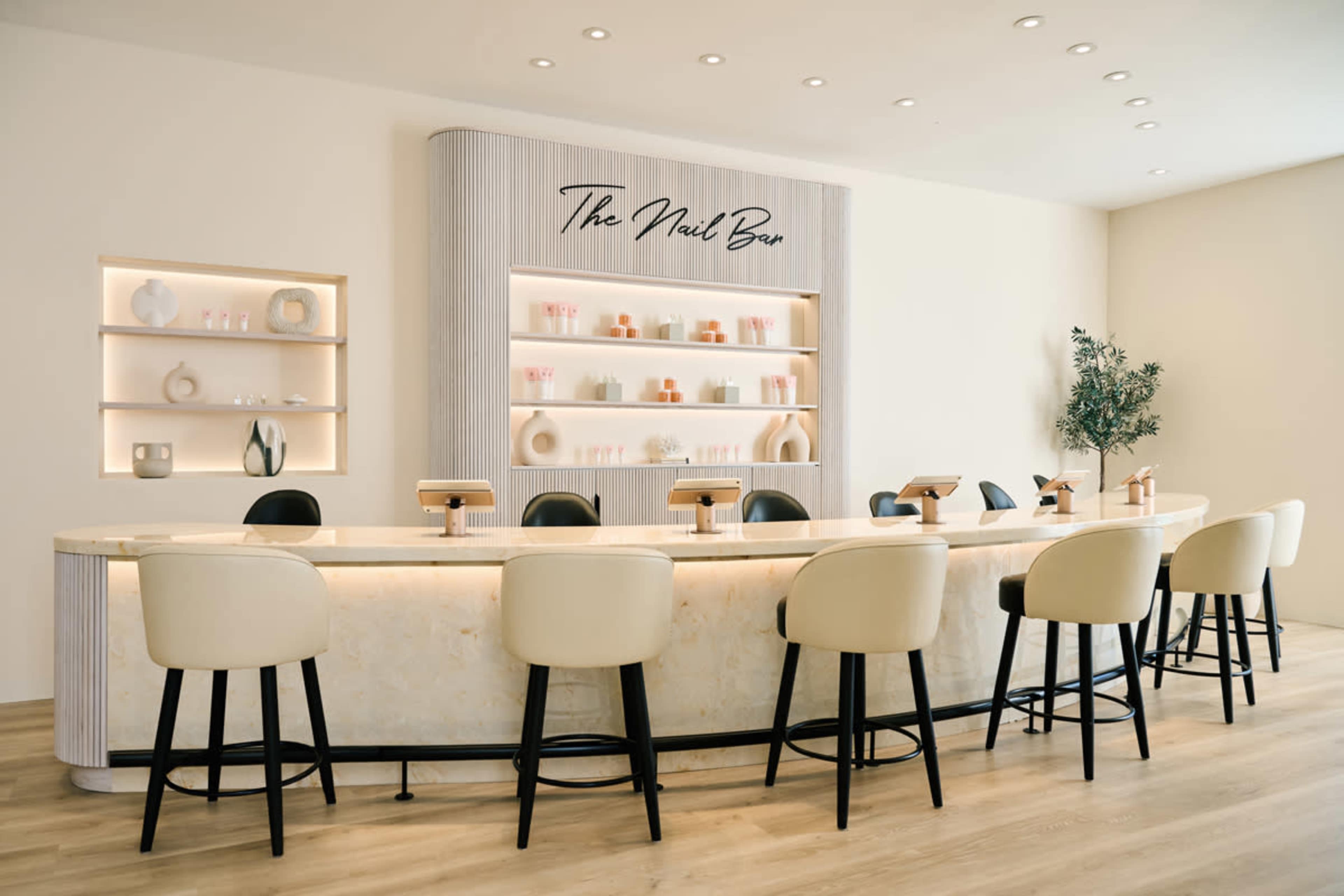 The image shows a modern nail bar with a curved marble counter and high-backed cream-colored stools, set against a light, minimalist backdrop.