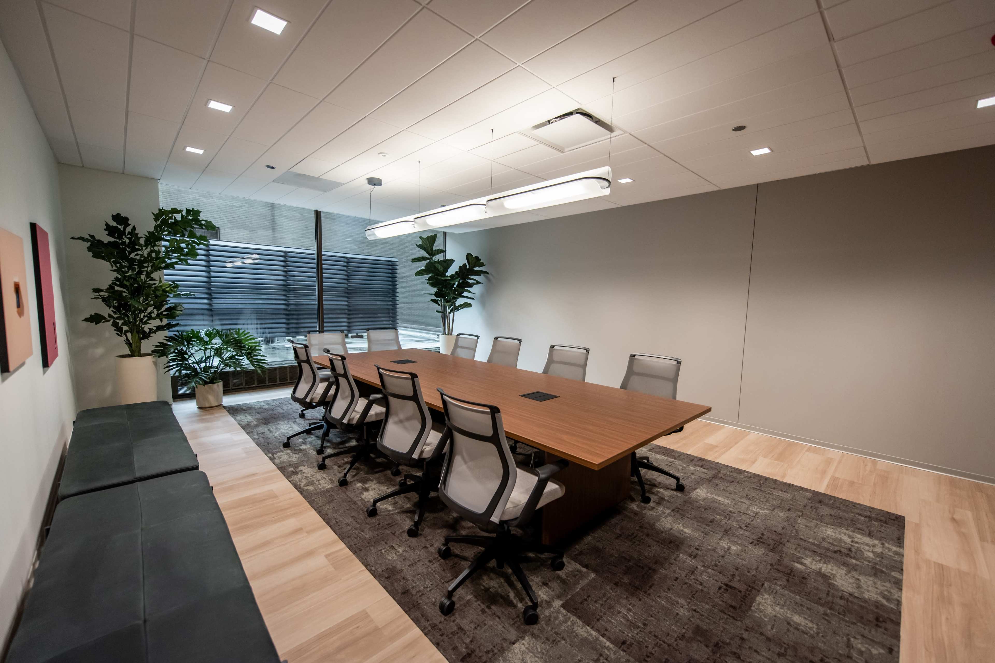 A modern conference room features a large wooden table surrounded by ergonomic chairs, with plants and large windows along one wall.