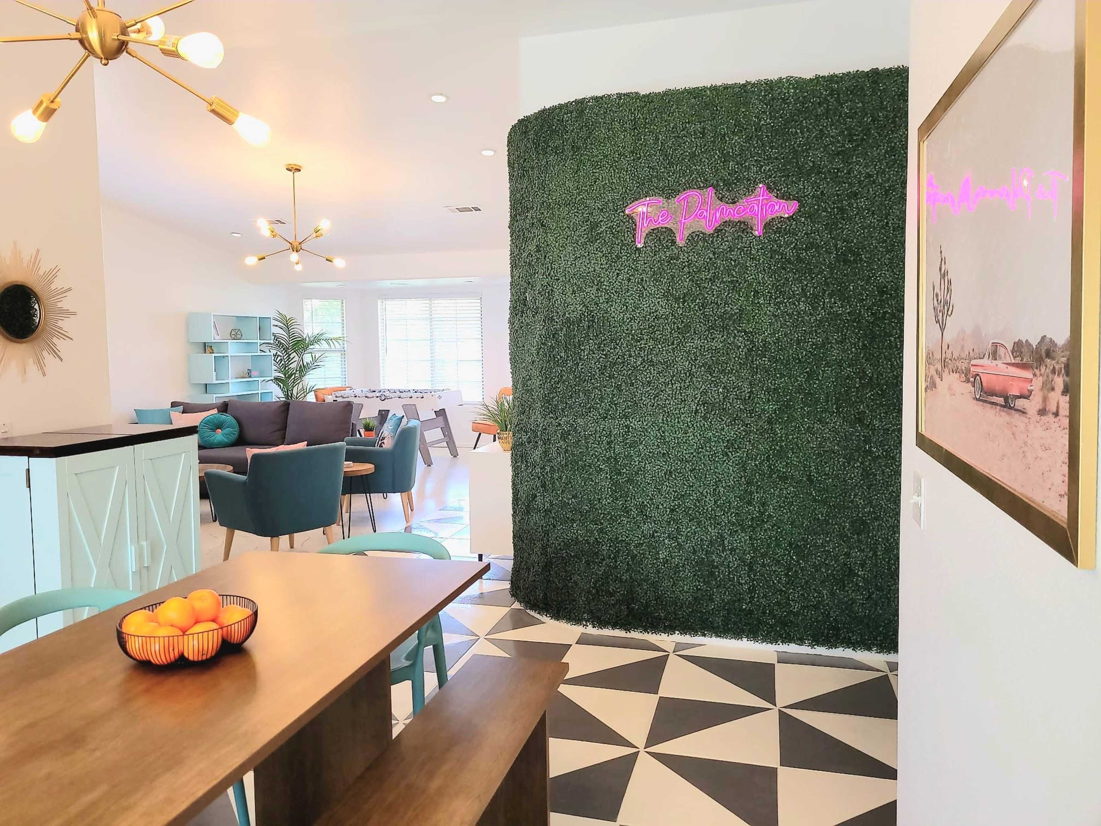 The image shows a vibrant interior space featuring a green wall with neon signage, a dining table with a fruit bowl, and modern furniture arranged in a living area.