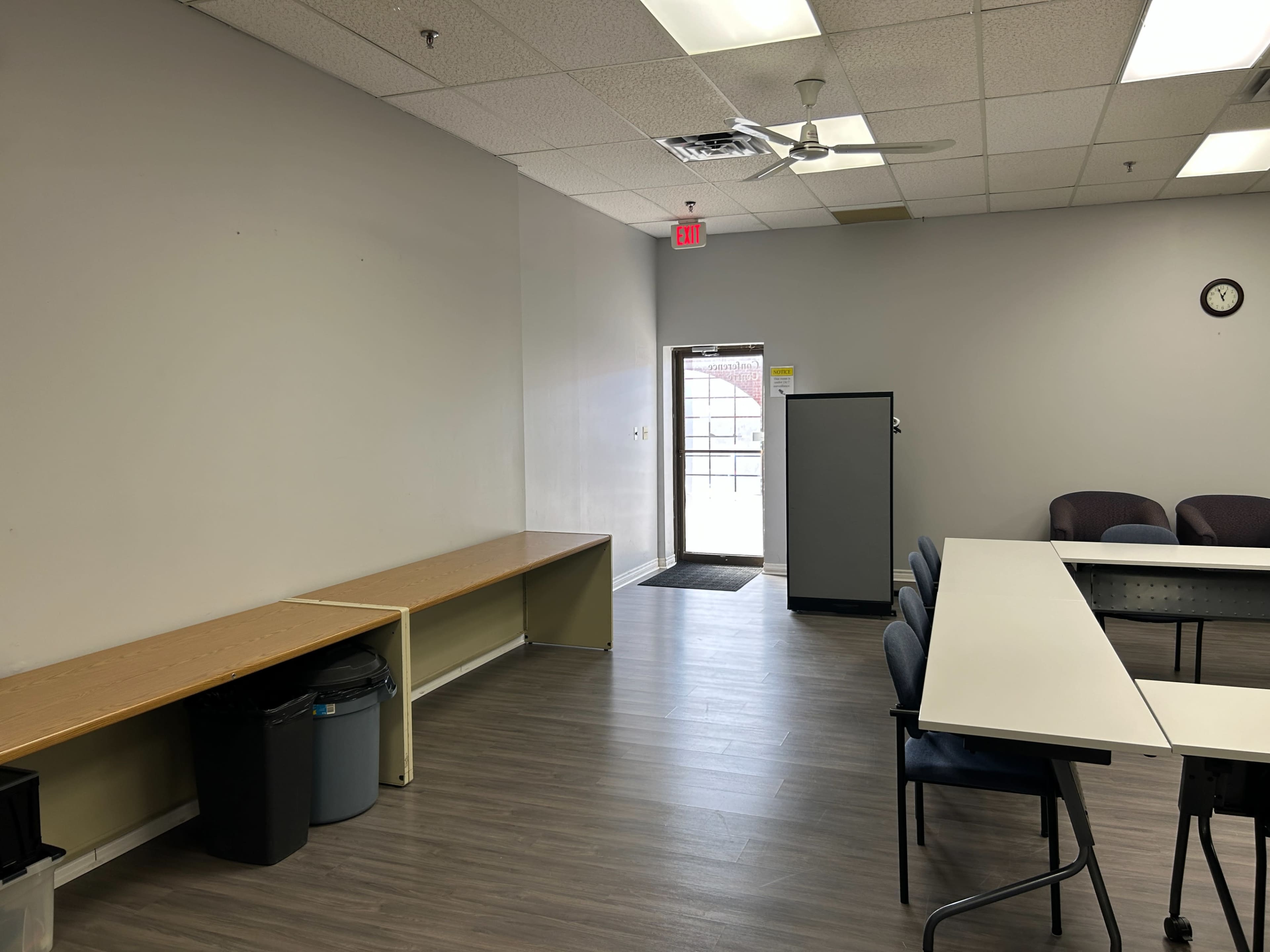 The image shows a sparsely furnished room with a long table, chairs, a trash can, and a door leading outside.