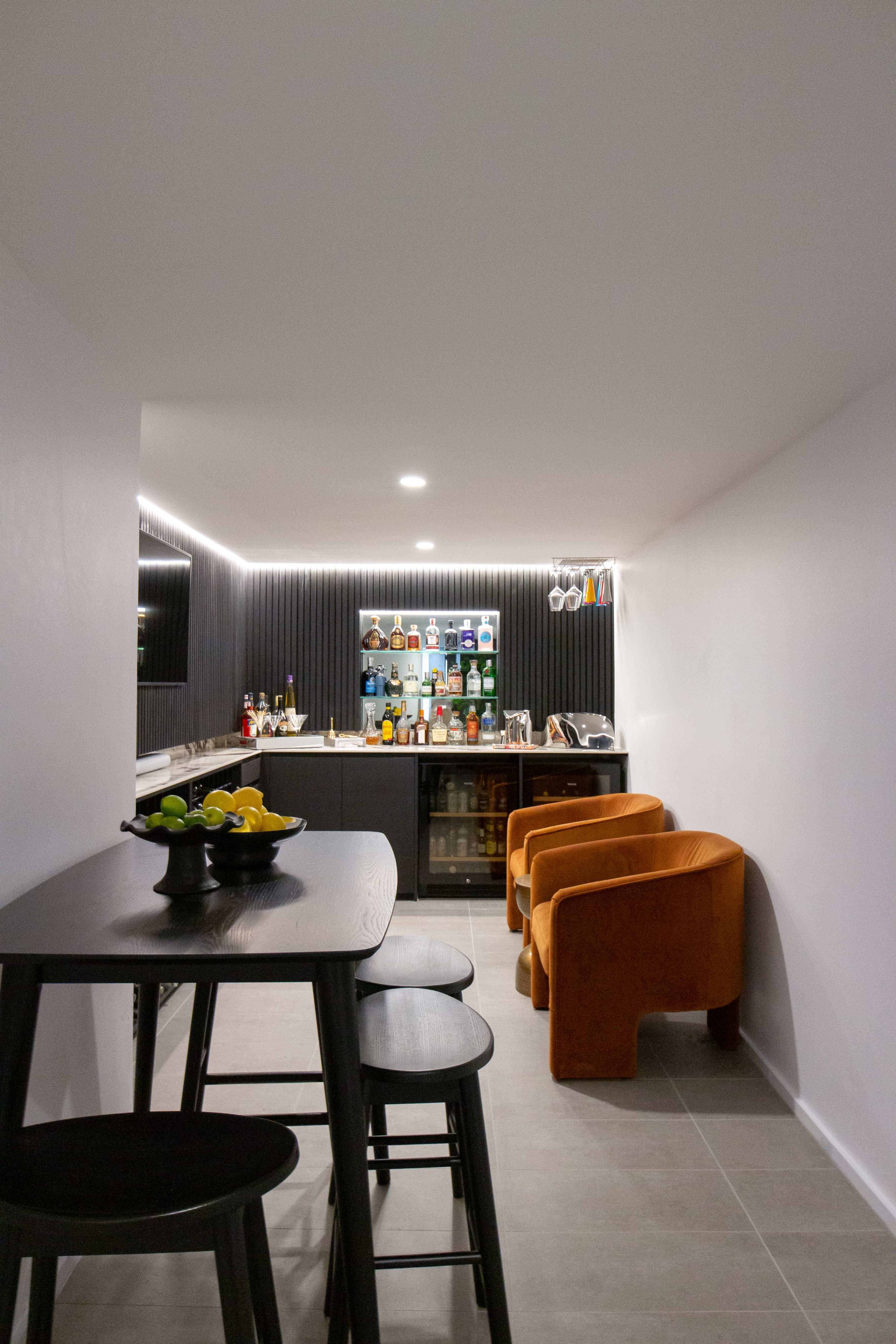 A modern home bar area with a black countertop, various bottles displayed on shelves, and orange chairs next to a black dining table.