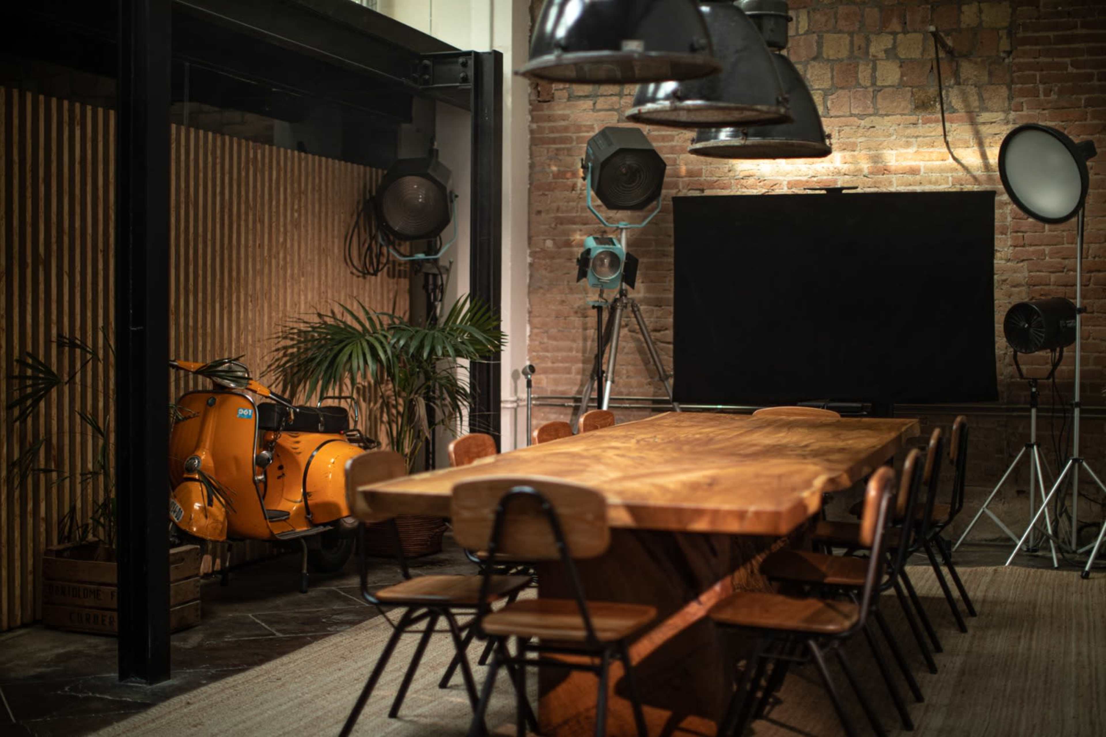 A large wooden dining table surrounded by black chairs is positioned in a room with industrial lighting, a vintage orange scooter, and studio equipment in the background.