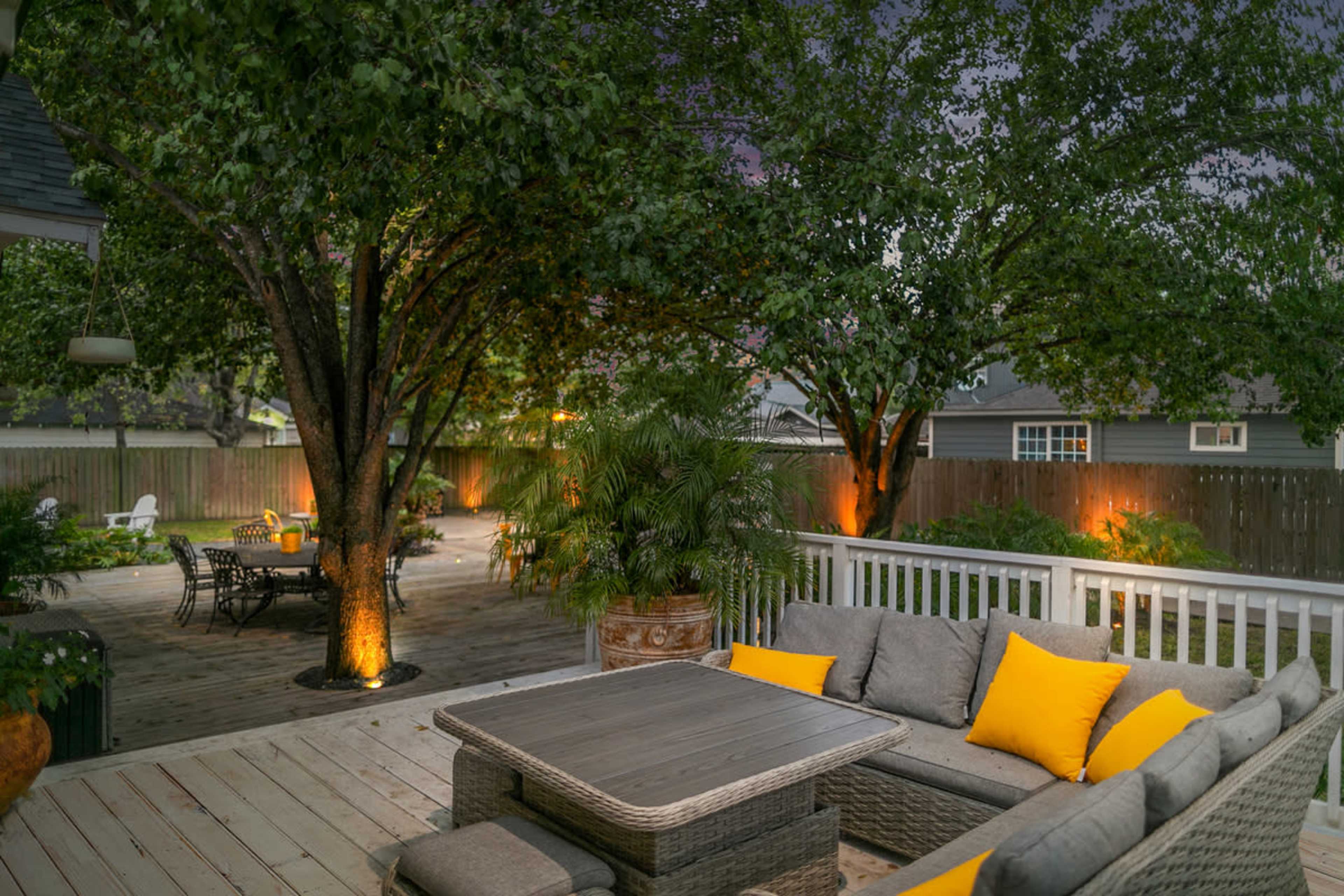 A backyard patio with a seating area and a dining set surrounded by trees and softly illuminated landscaping.